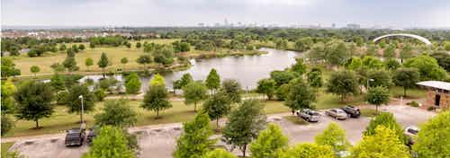 Overview of scattered vibrant green trees under a blue sky surrounding a grassy lake area with cars parked in a nearby lot