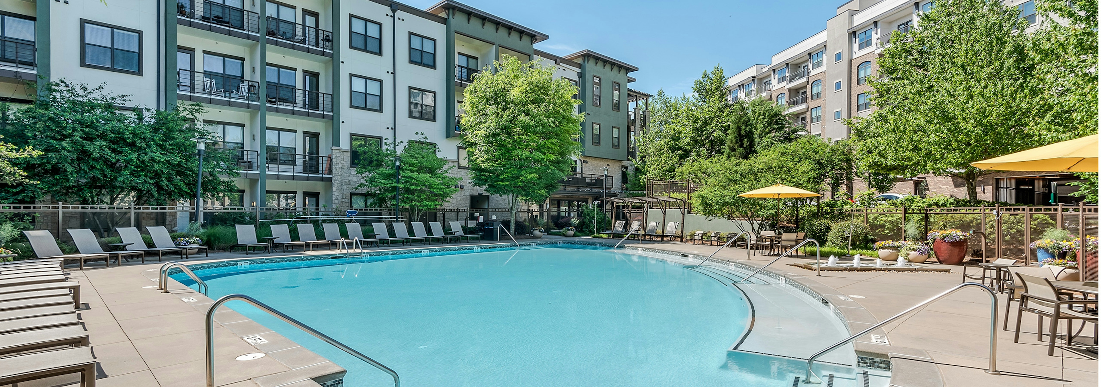 Exterior of pool at AMLI Parkside surrounded by lounge chairs with building facade and trees in the background on a sunny day