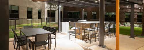 Nighttime view of barbecue area at AMLI on Aldrich with outdoor tables and chairs under covering and surrounded by shrubbery
