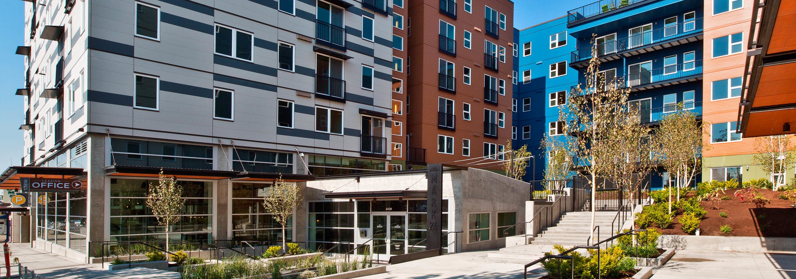 Exterior entrance to AMLI Mark24 white blue and orange building with patios and trees bushes and view of the sidewalk