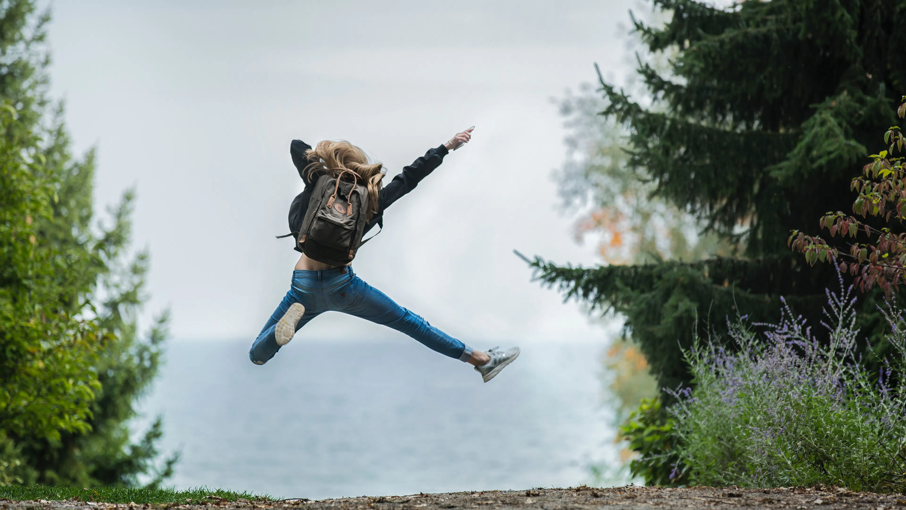 woman on a hiking trail jumping into the air pointing to the sky surrounded by large trees