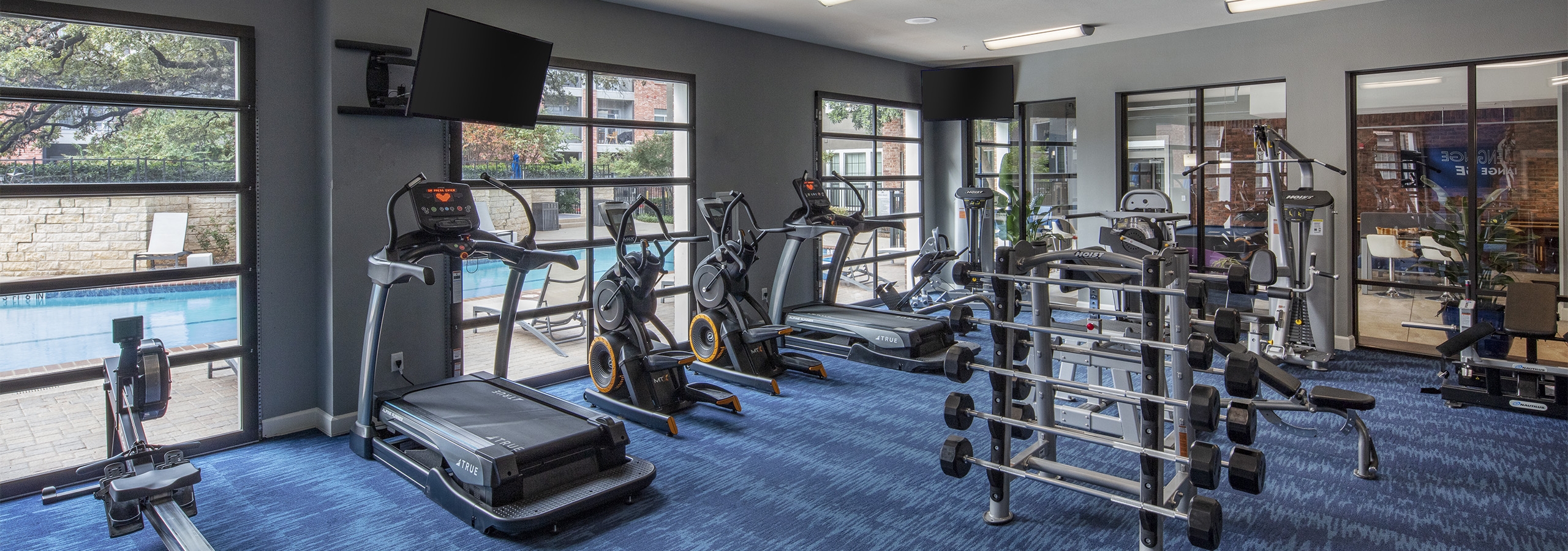 Day view of AMLI Eastside poolside fitness center with a cardio and strength training machines and TVs set on blue carpet