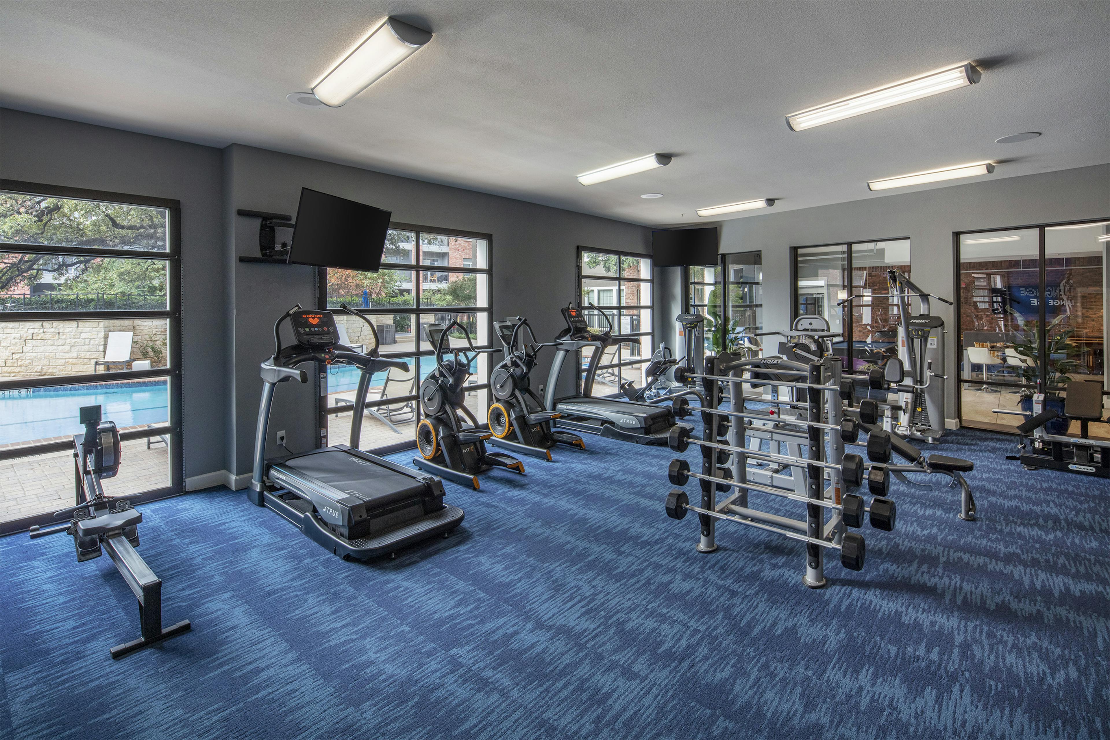 Day view of AMLI Eastside poolside fitness center with a cardio and strength training machines and TVs set on blue carpet