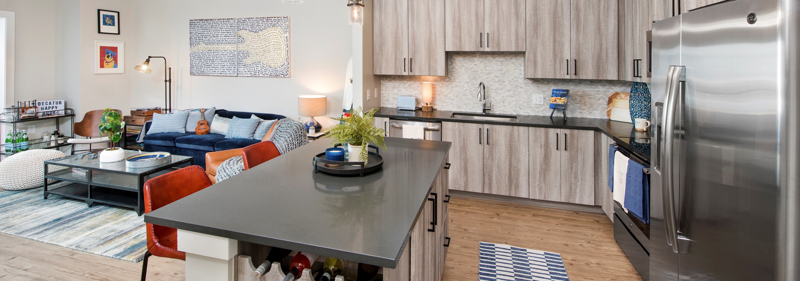 AMLI Decatur kitchen with dark counters and light wood cabinets with stainless steel appliances and living room in background