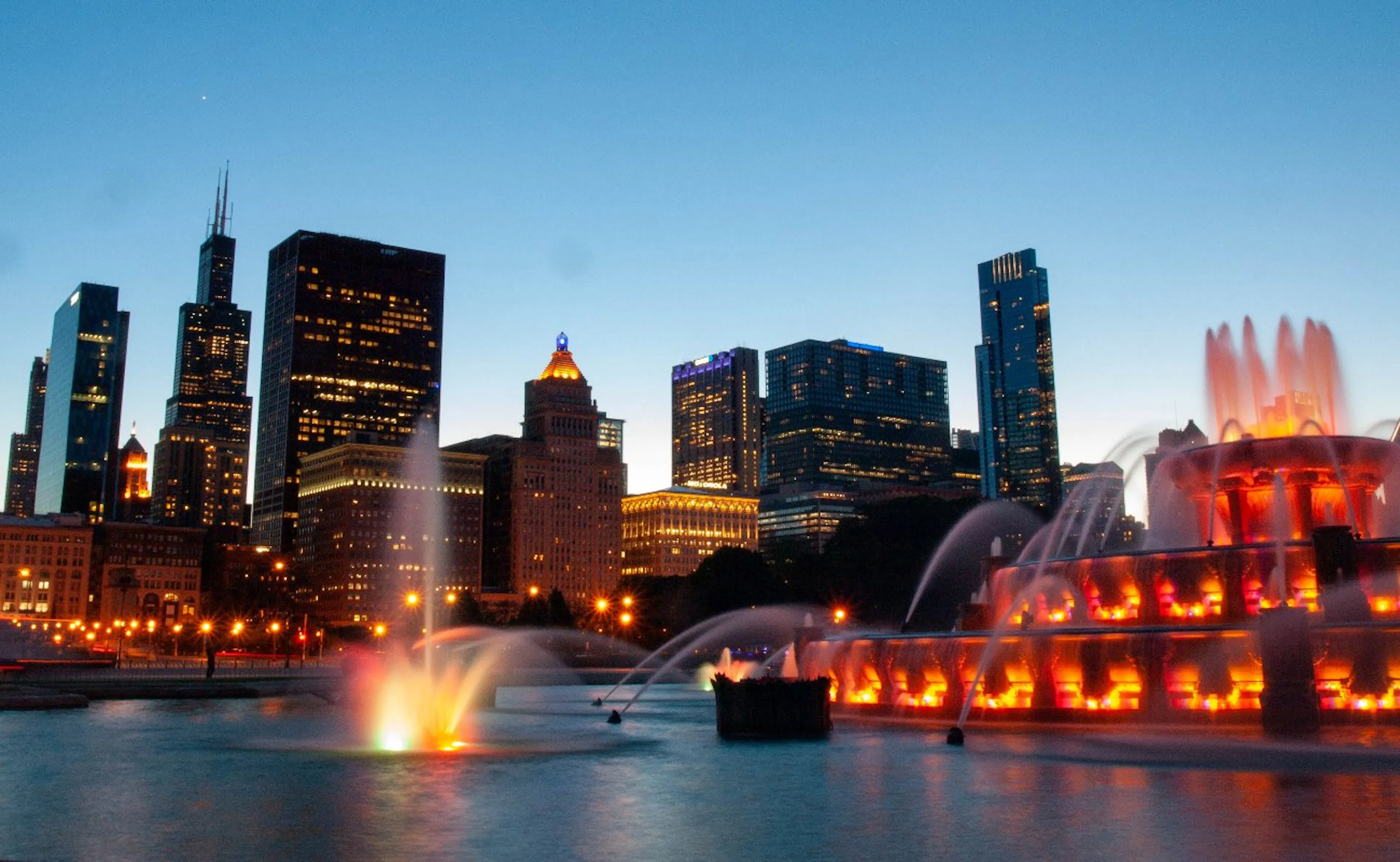 View of Buckingham Fountain in Grant Park lit up at night against the Chicago skyline with the Sears Tower showing