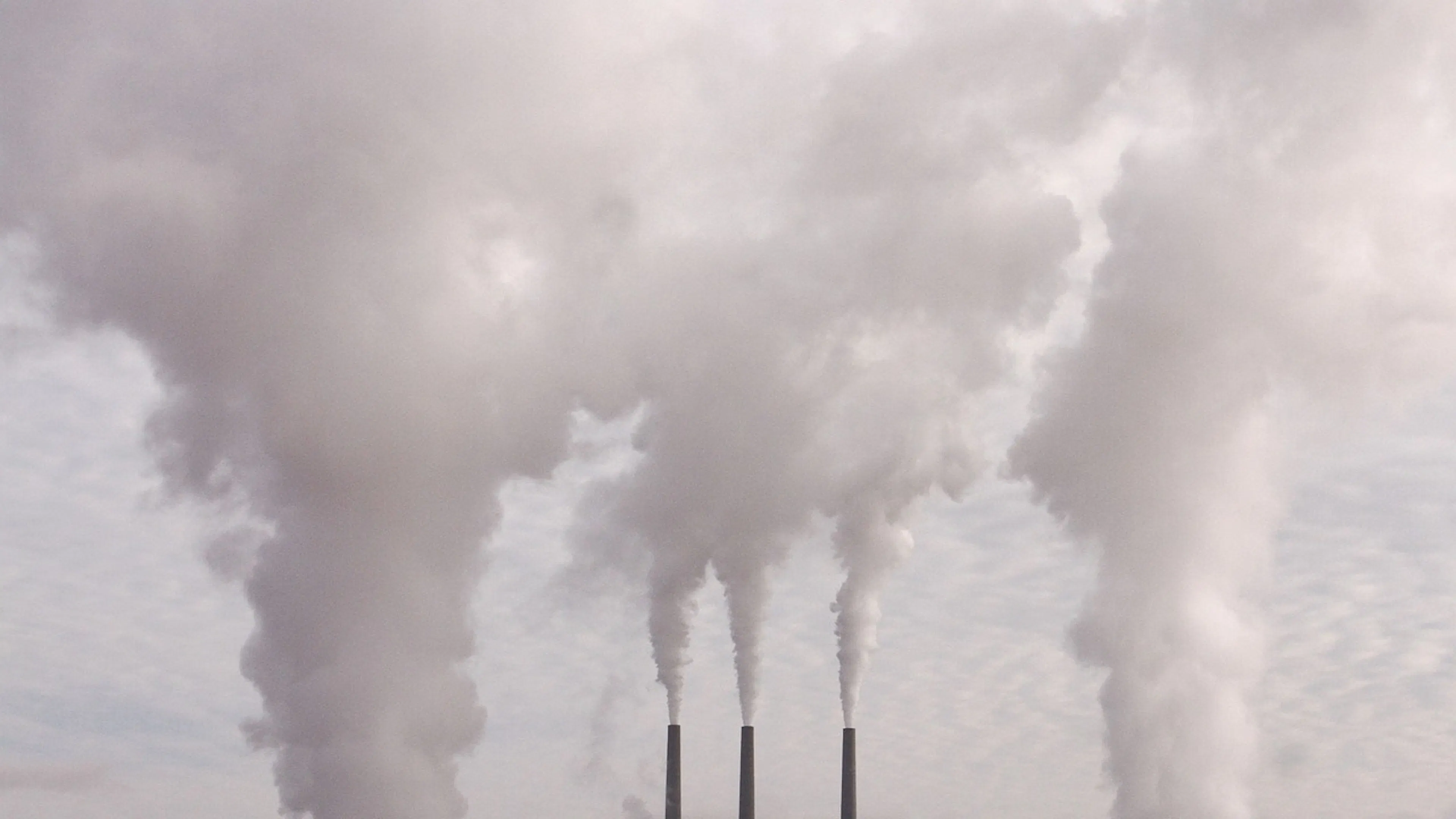 Three tall smokestacks billowing steam and smoke out from a small factory in the middle of a large field