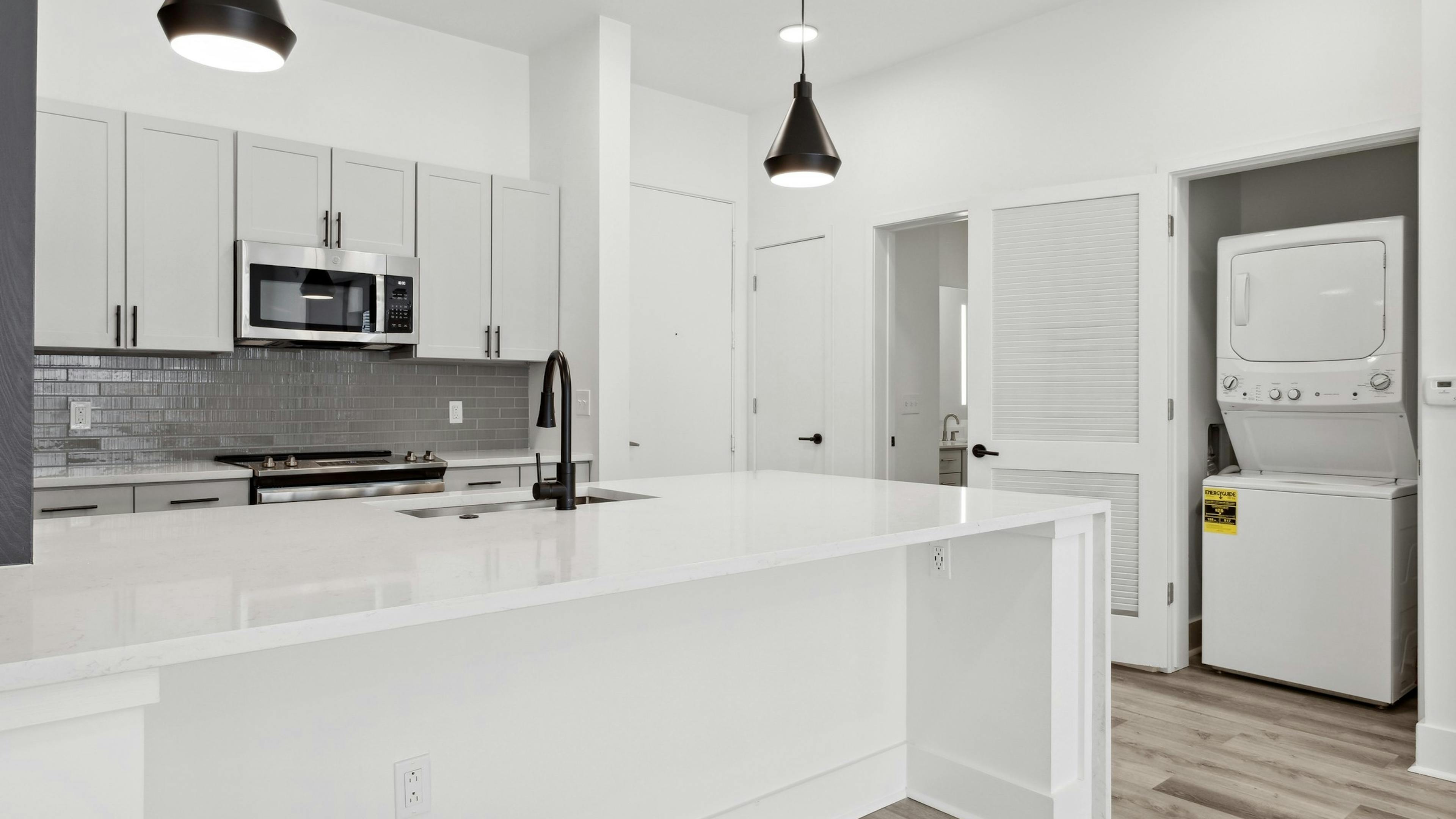 Island kitchen with white counter tops and cabinets and gray tiled backsplash with view of laundry closet at AMLI Old 4th Ward