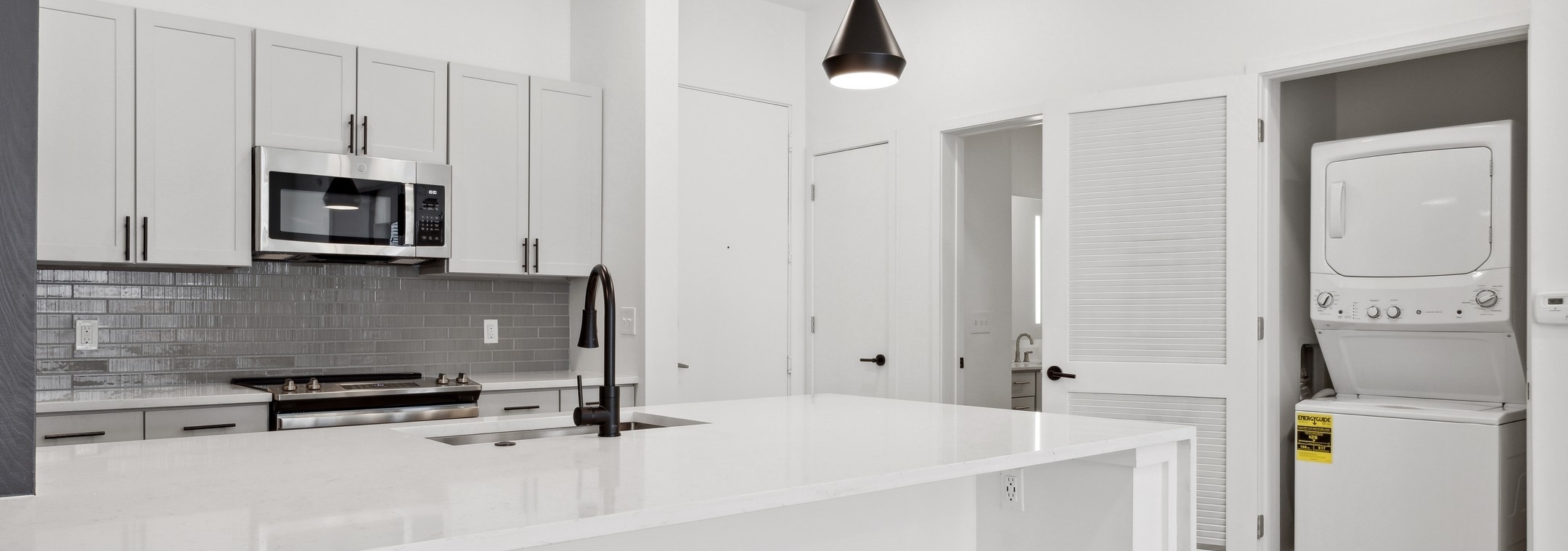 Island kitchen with white counter tops and cabinets and gray tiled backsplash with view of laundry closet at AMLI Old 4th Ward