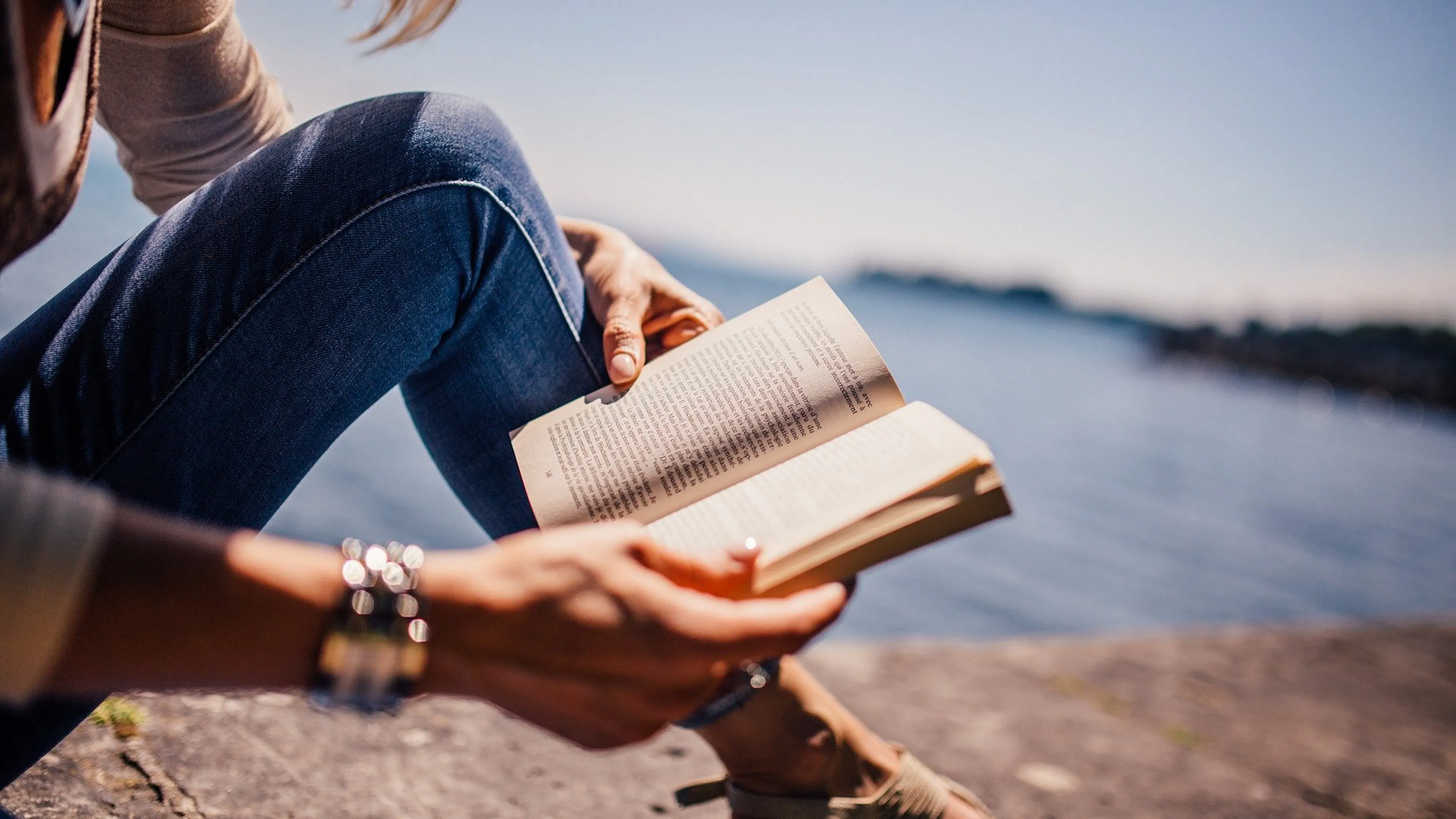 A woman holding a book and reading by a lake on a sunny day