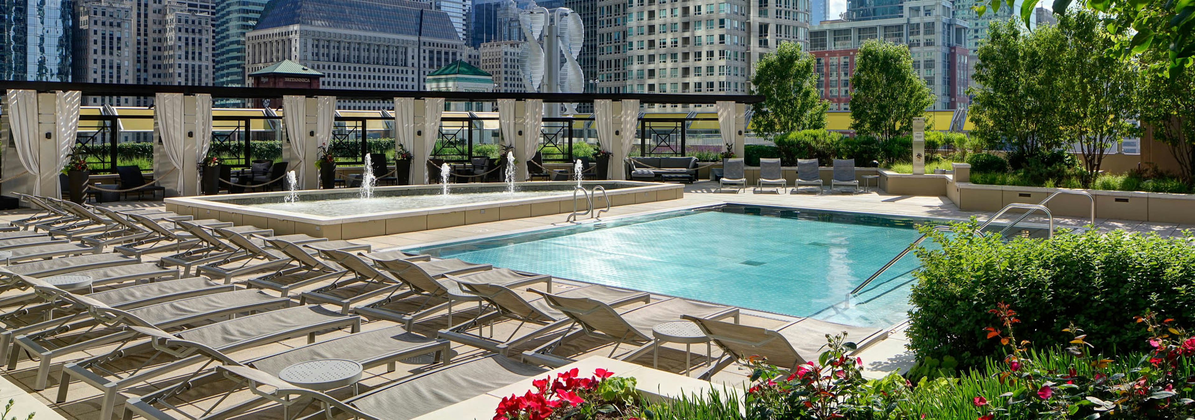 View of the pool at AMLI River North with surrounding beige lounge chairs and vibrant foliage with a city skyline in the back