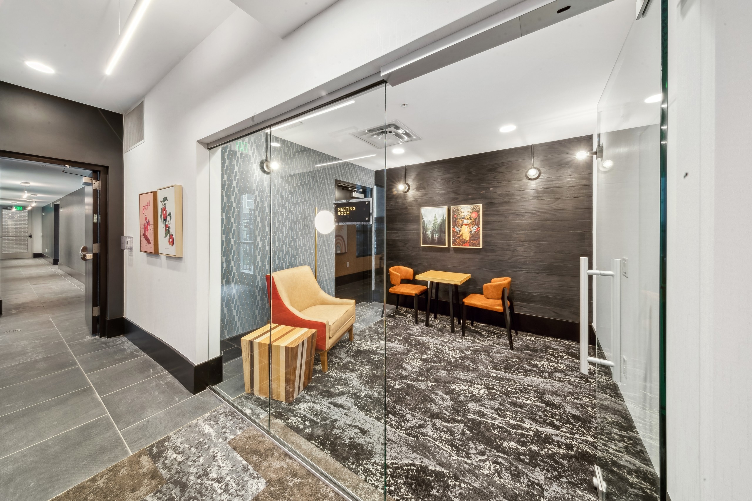 AMLI RiNo business center meeting room with a glass door and guest chair and wood table with 2 chairs on dark paneled wall 
