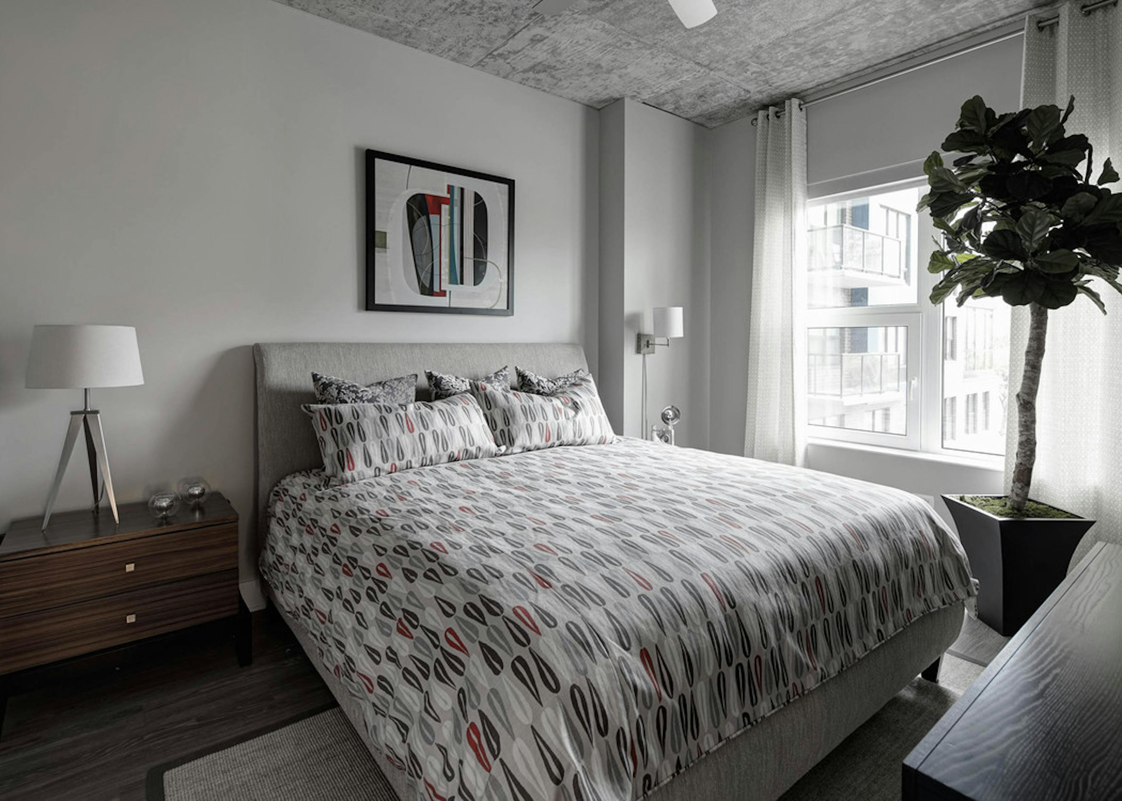 A simplistic bedroom at AMLI Lofts decorated in earth tones with a large faux tree next to a window with a bright city view