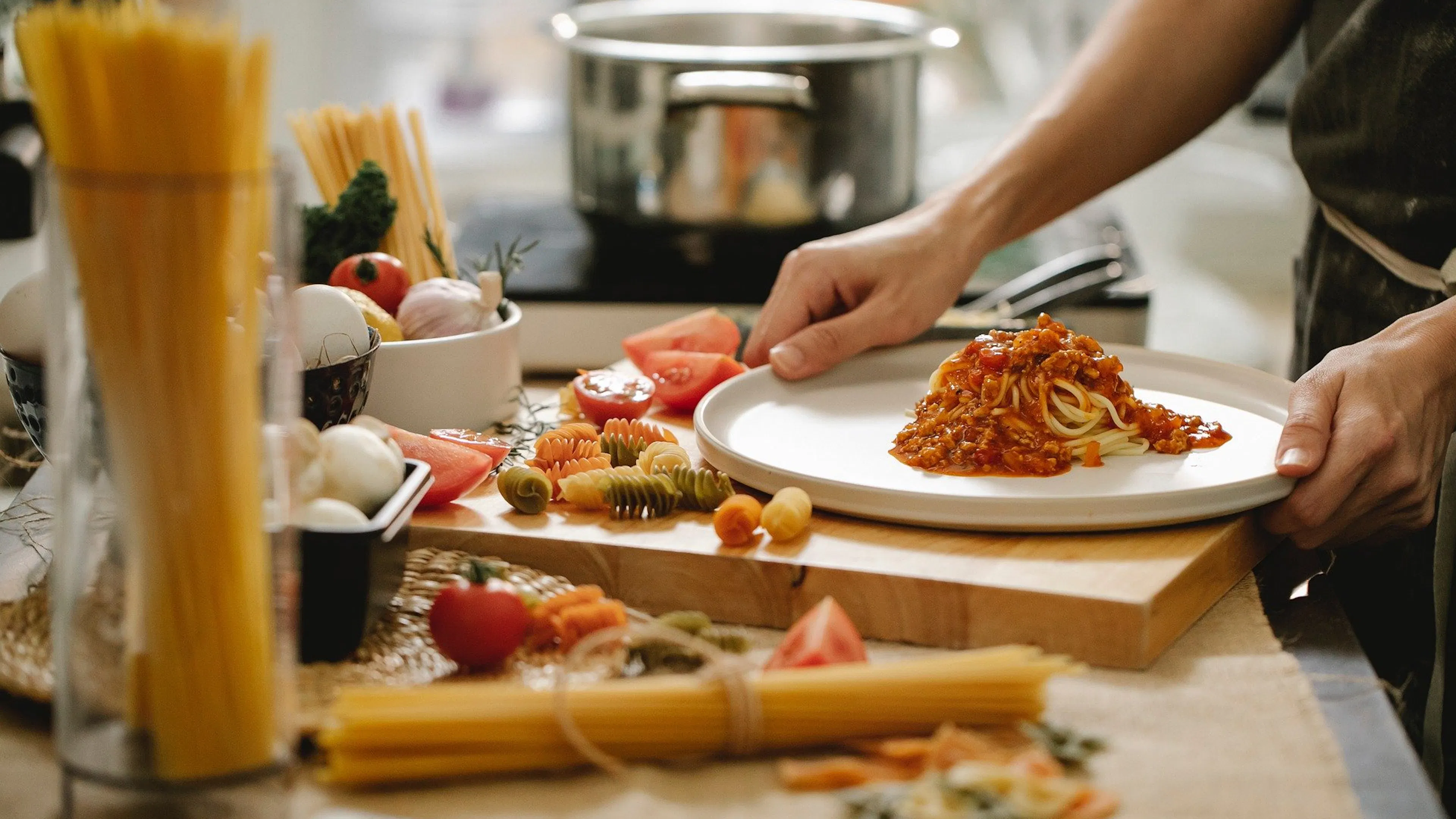 A person cooking pasta with fresh vegetables and spaghetti scattered all around