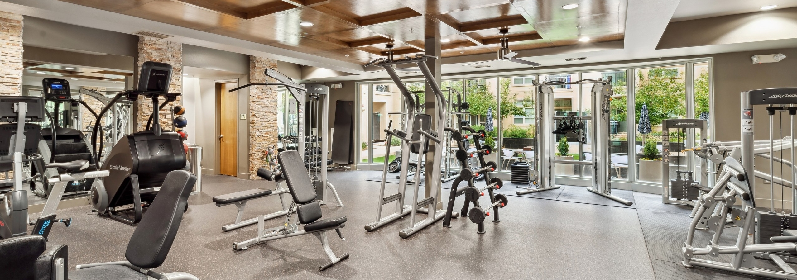 A fitness center at AMLI RidgeGate with free weights and a bench press and a mirror and a view into the community courtyard