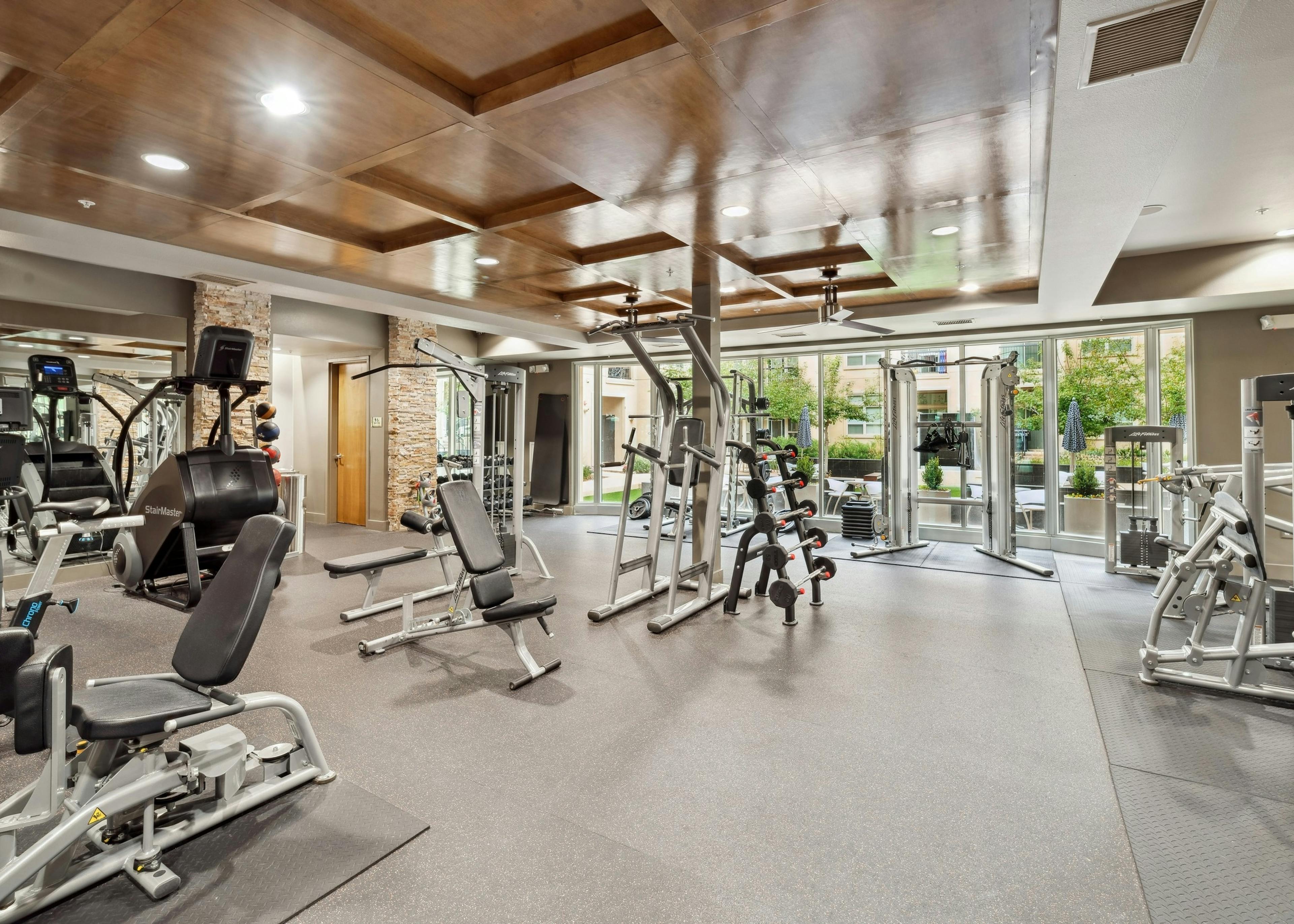 A fitness center at AMLI RidgeGate with free weights and a bench press and a mirror and a view into the community courtyard