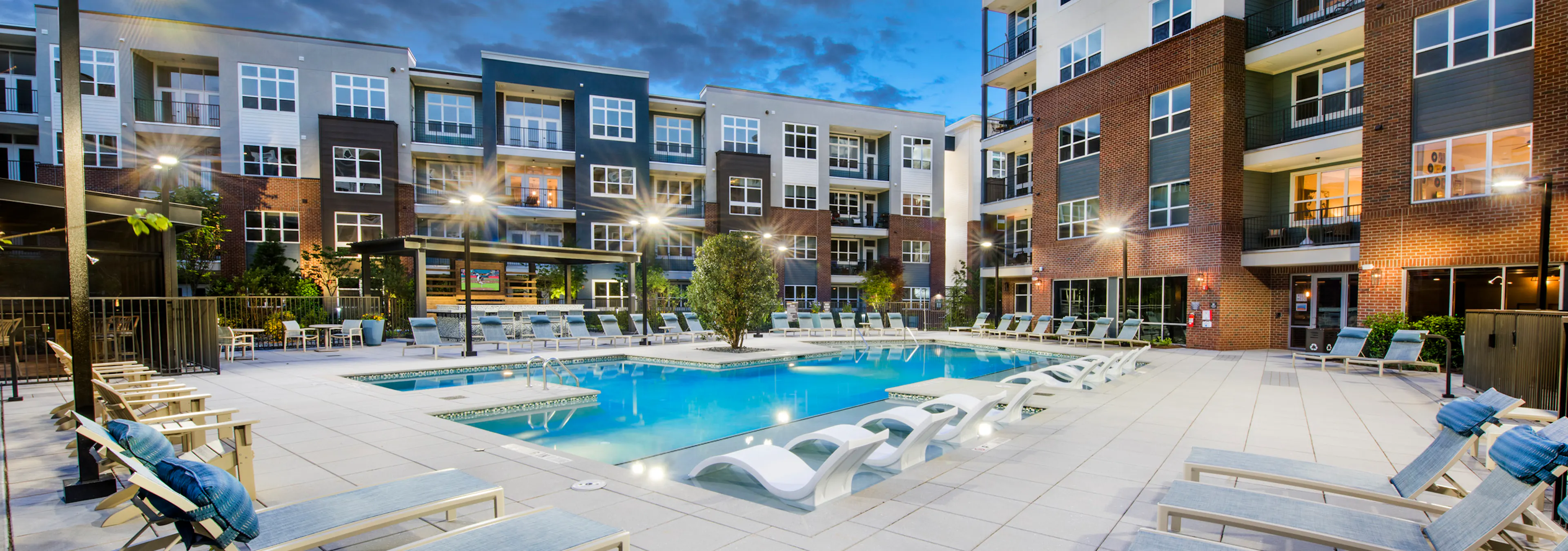 Evening view of AMLI Decatur pool with potted plants and blue water and white lounge chairs with blue pillows around the pool