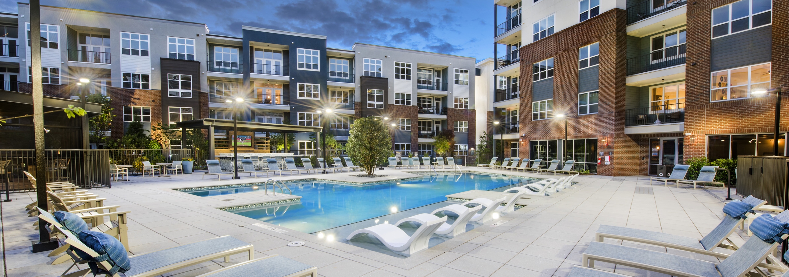 Evening view of AMLI Decatur pool with potted plants and blue water and white lounge chairs with blue pillows around the pool