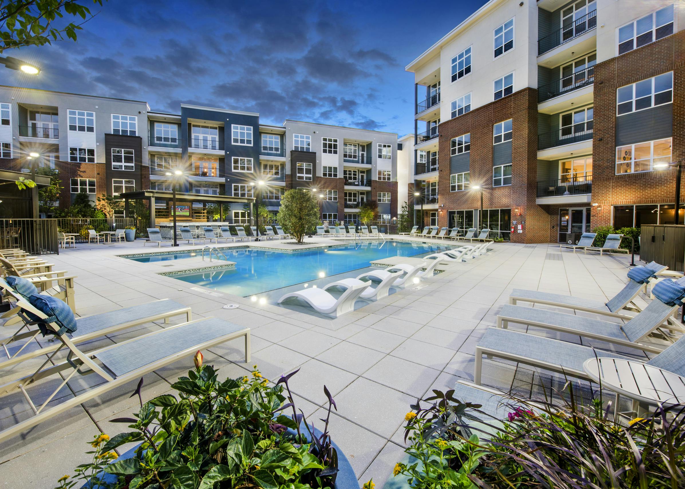 Evening view of AMLI Decatur pool with potted plants and blue water and white lounge chairs with blue pillows around the pool