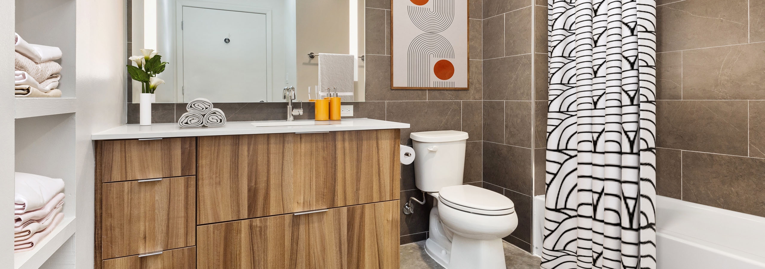 View of bathroom with wood vanity with large mirror above and white toilet and bathtub with geometric curtain at AMLI on 2nd