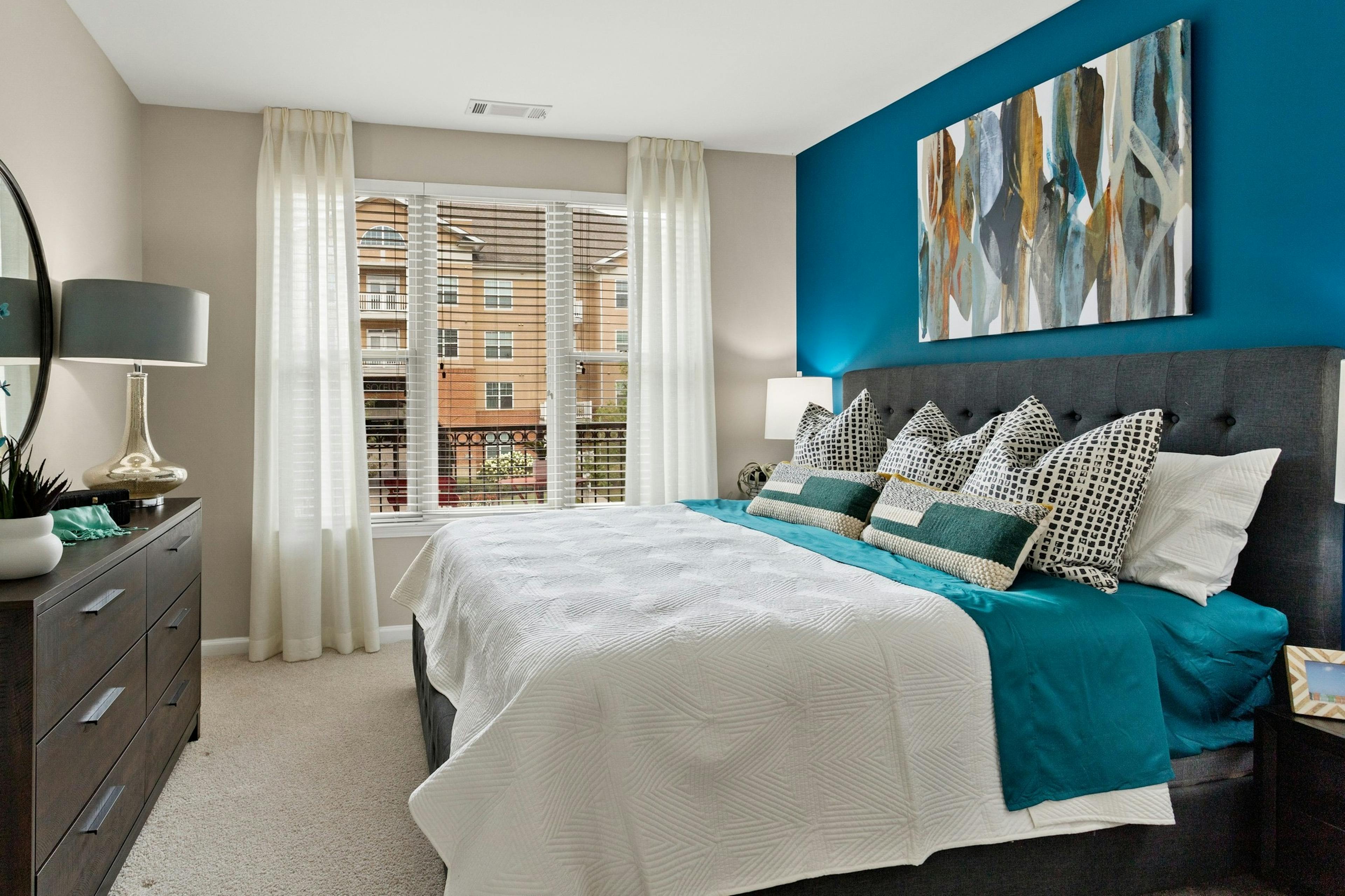 Bedroom at AMLI Lindbergh apartments with king bed on blue wall and dark wood dresser and white curtains over large window