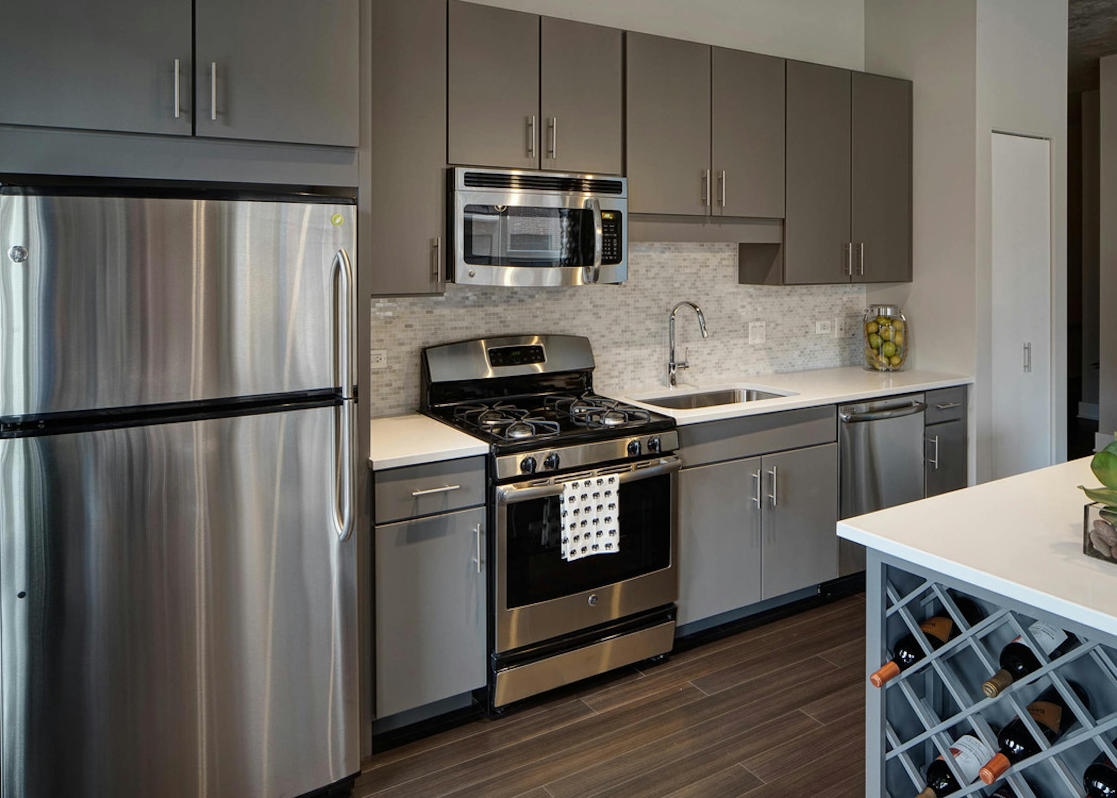 An AMLI Lofts kitchen containing stainless steel appliances and grey cabinets paired with quartz countertops and a backsplash
