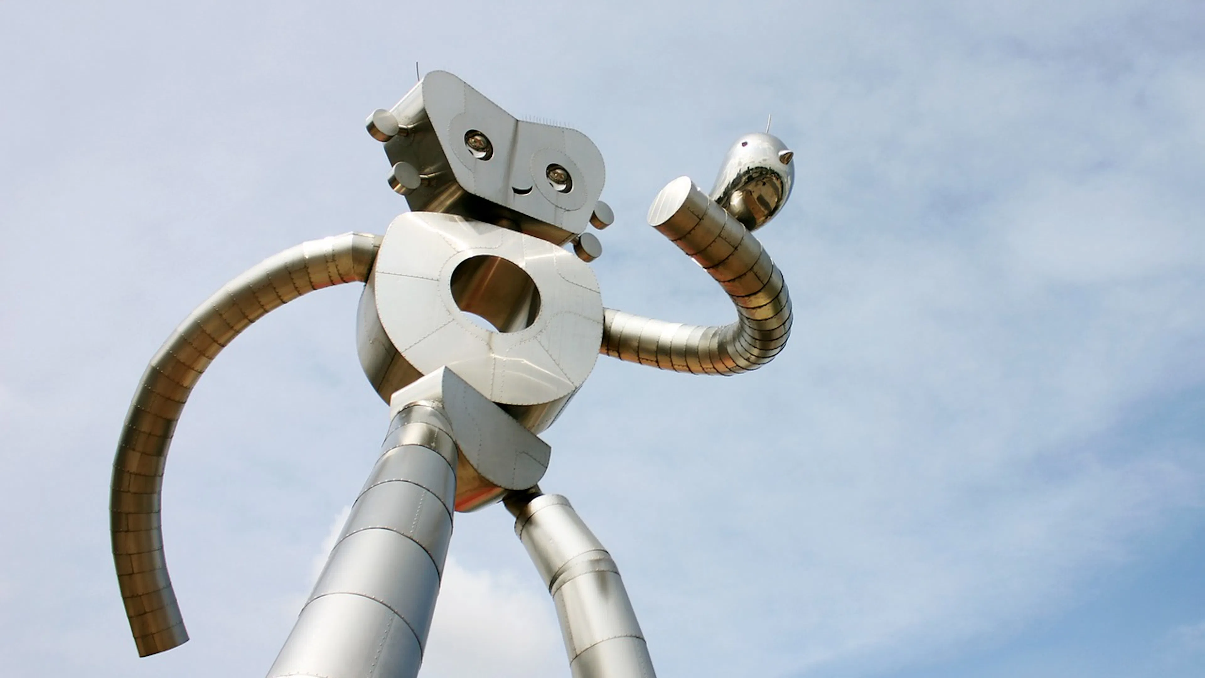 A large metal robot sculpture smiling and holding a metal bird on its arm set against a blue sky and wispy clouds