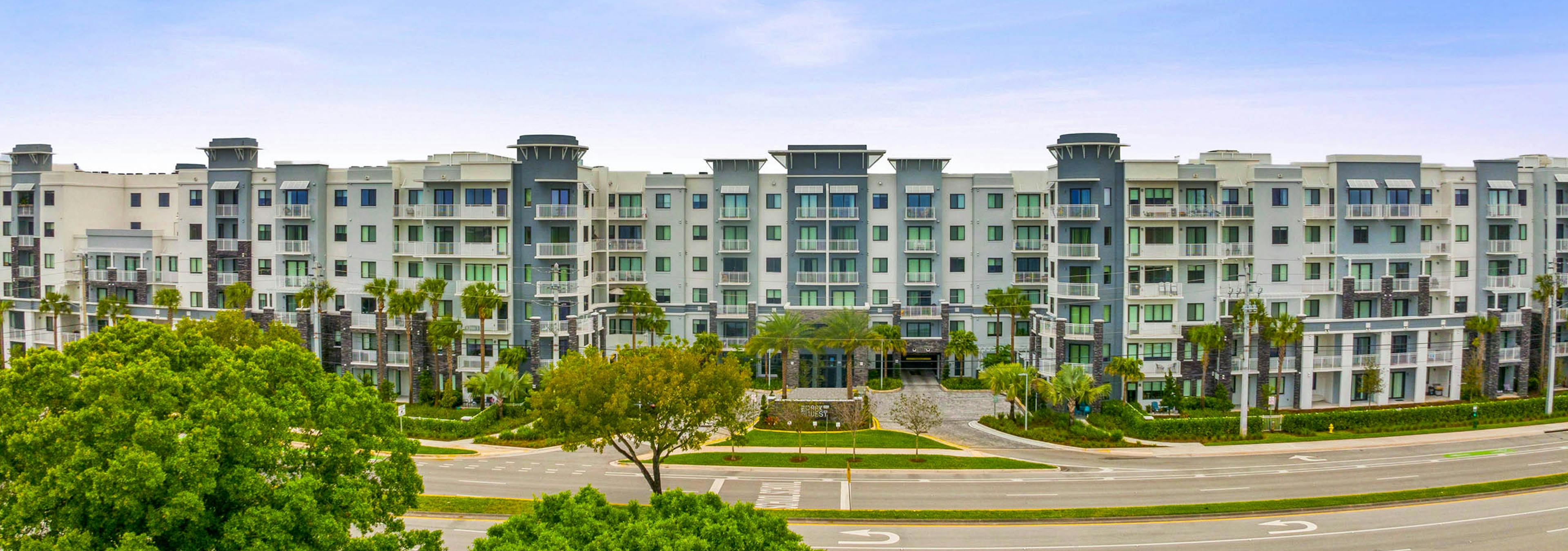 Distant view of AMLI Park West apartment building entrance with green trees all around and blue sky above