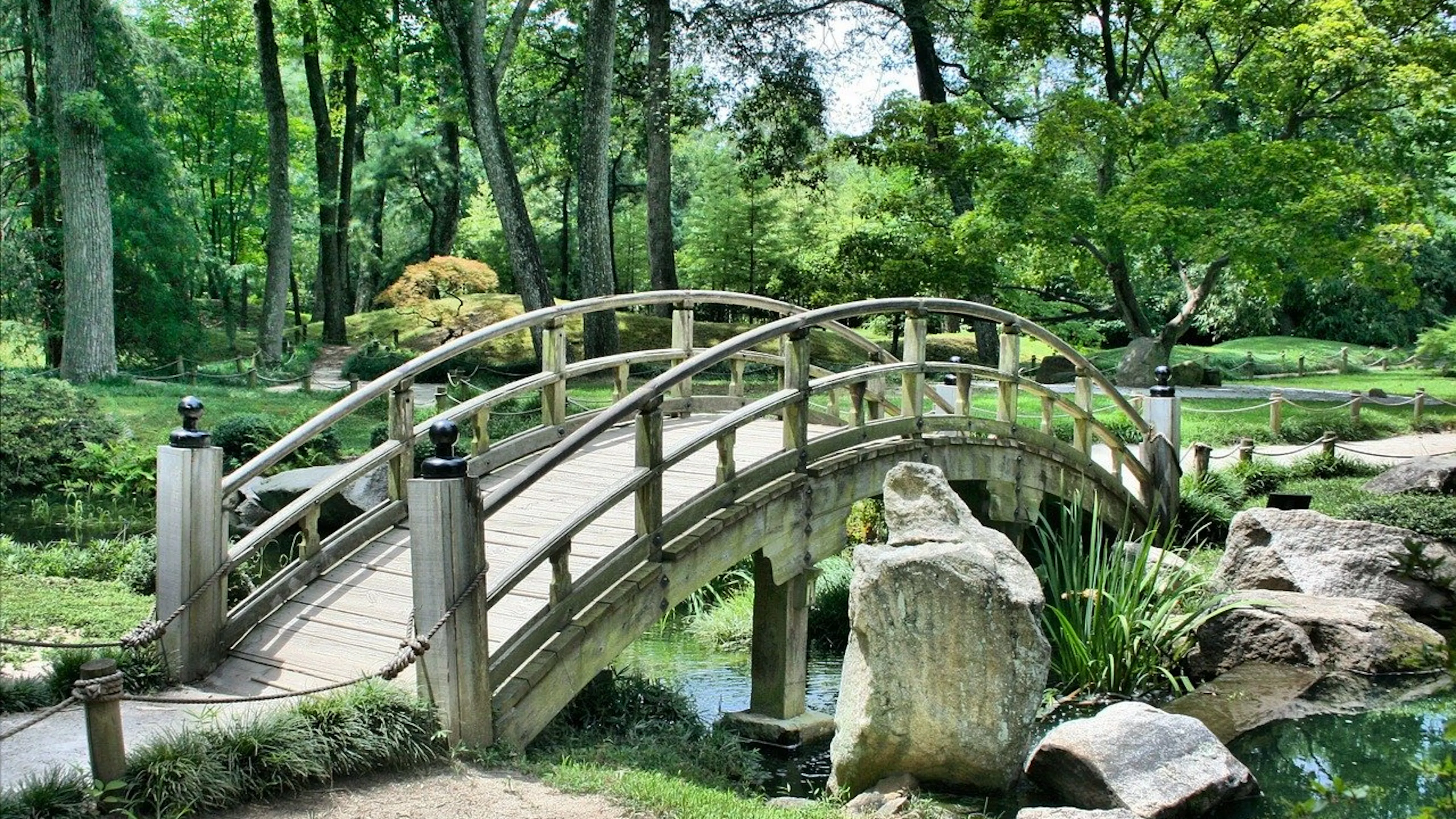 A short stone bridge crossing a little creek in a shady park surrounded by trees and grass on a sunny day