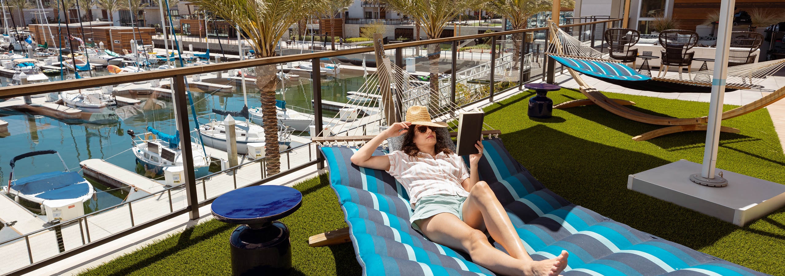 AMLI Marina Del Rey apartment resident laying on blue hammock reading a book with view of docked boats in marina