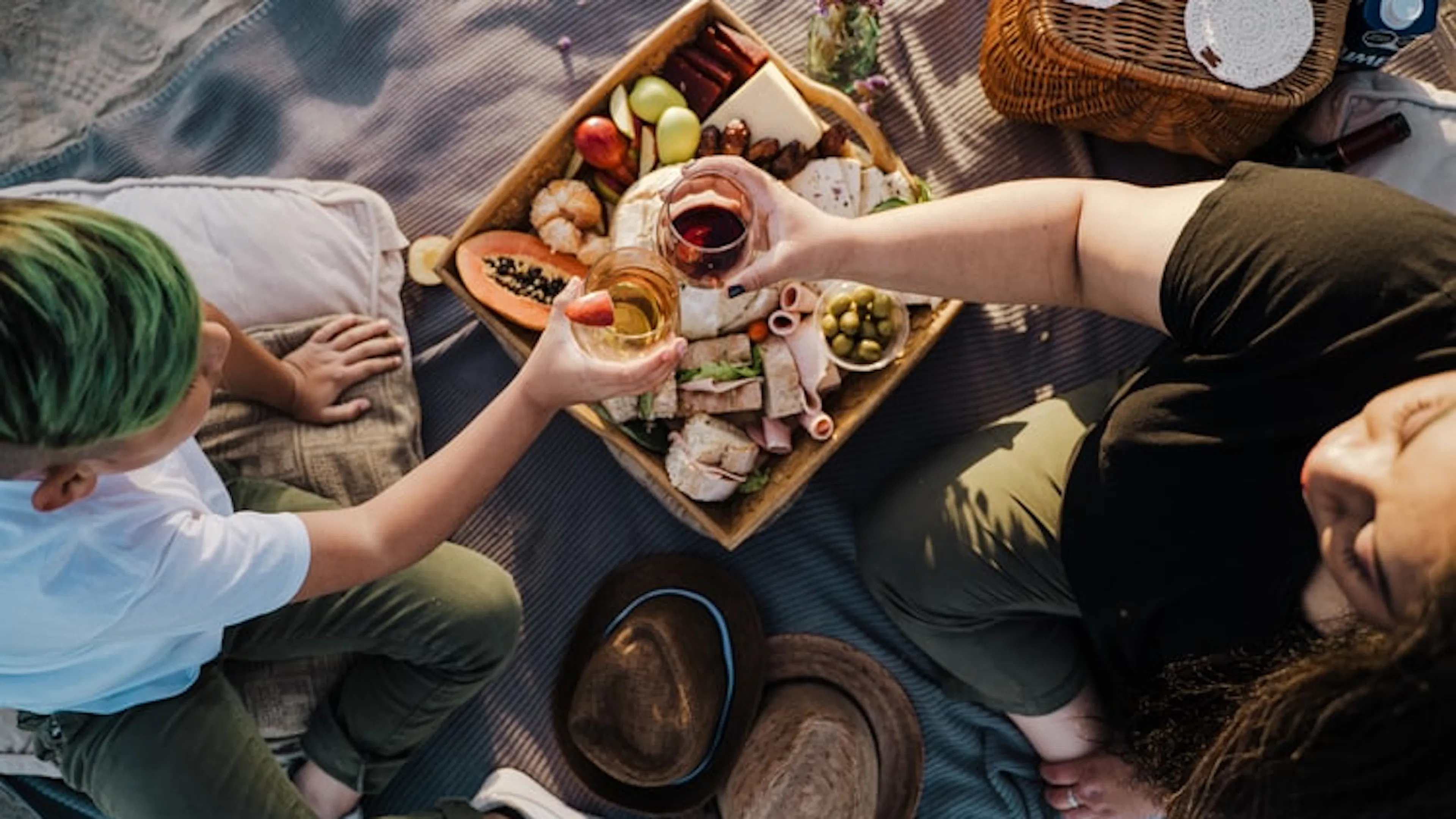 Two people on a gray picnic blanket drinking wine and eating fruit and cheeses
