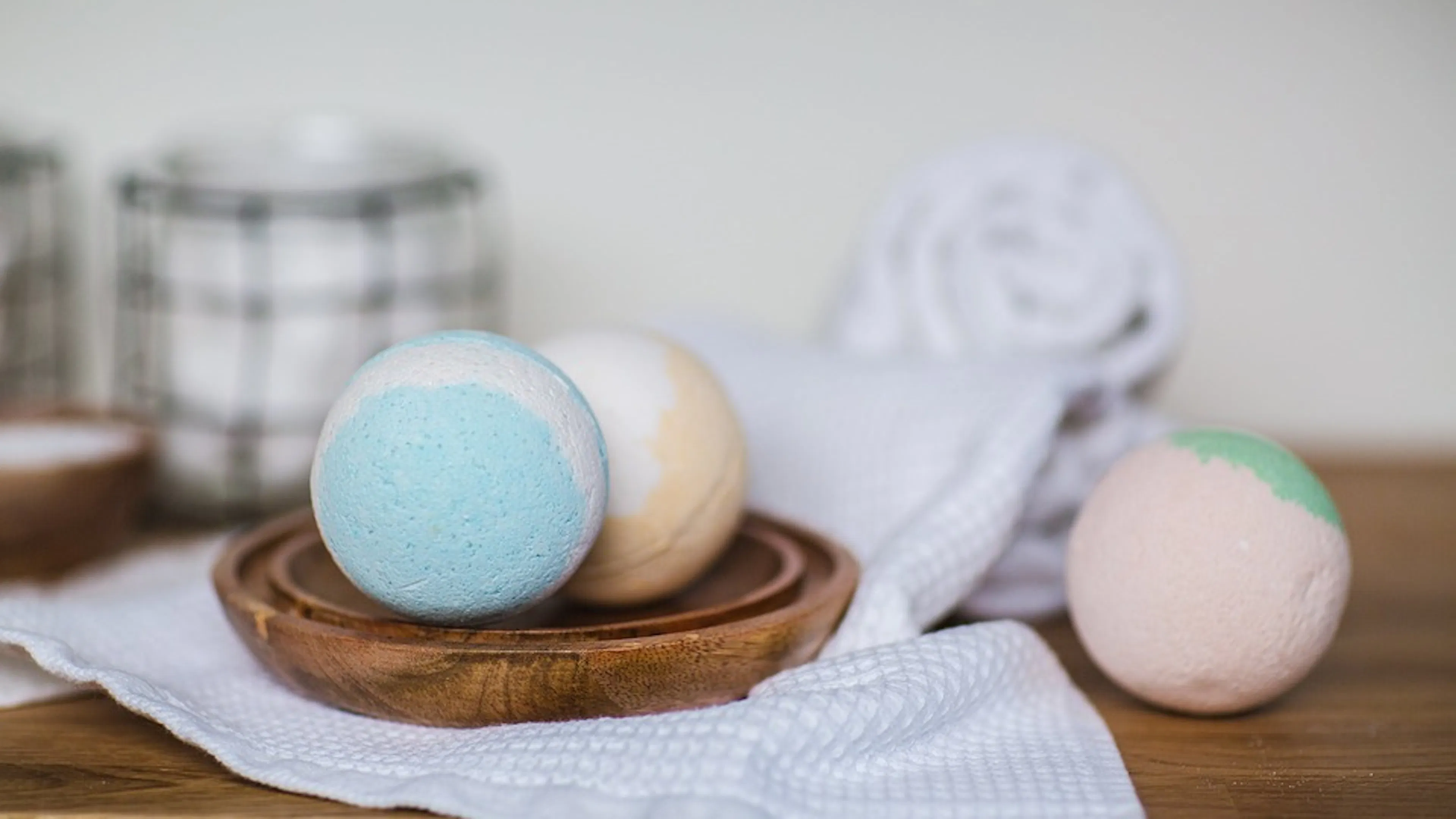 Three multi colored bath bombs on a wooden plate and light towel set on a wooden table