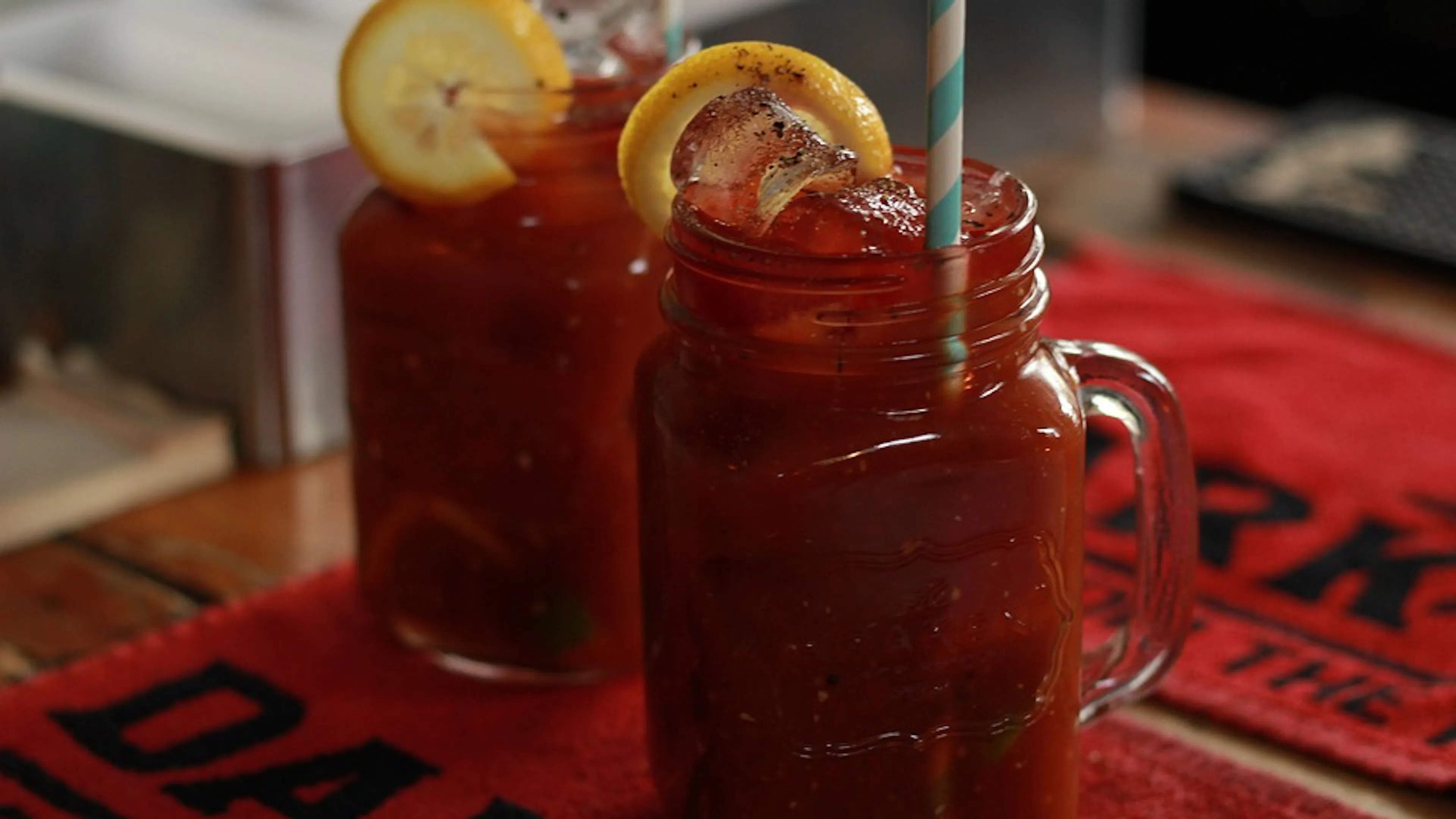 Two bloody Marys in glass Mason jars on a bar with lemon slices on their rims and a striped paper straw