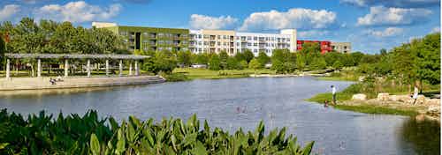 Lake with surrounding green foliage under a cloudy blue sky at Mueller Lake Park with AMLI at Mueller building in background