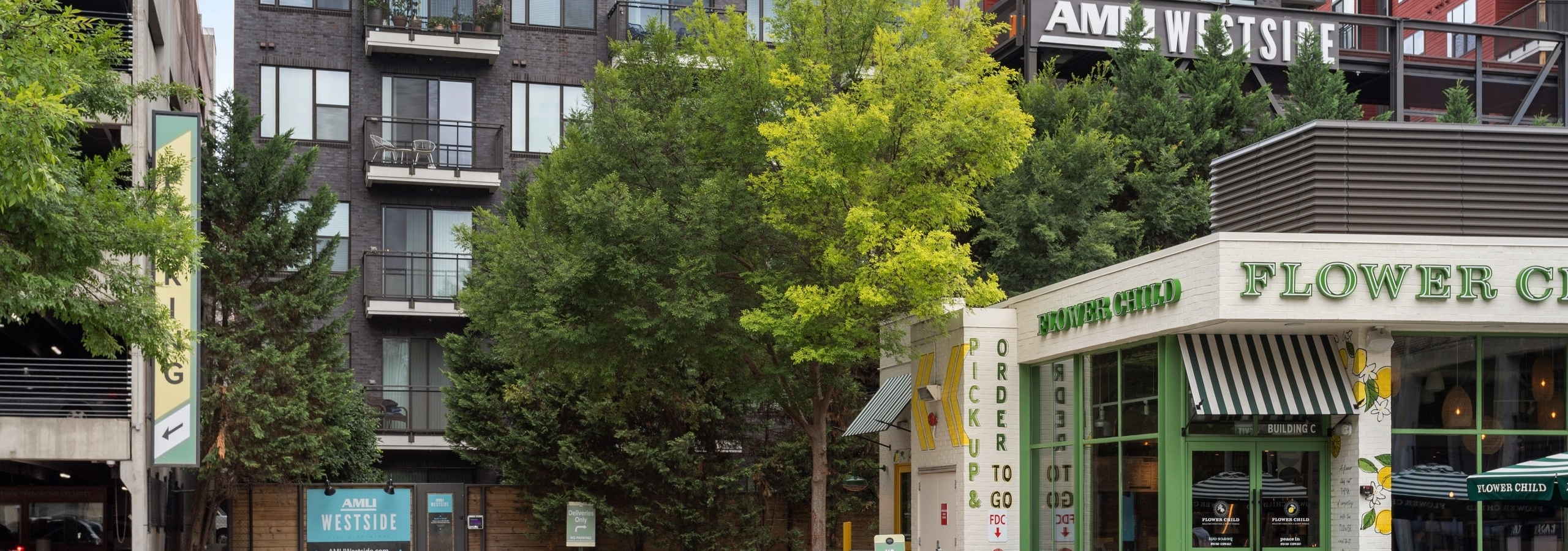 Daytime view of AMLI Westside apartment building's brick façade and monument sign and retail space surrounded by trees