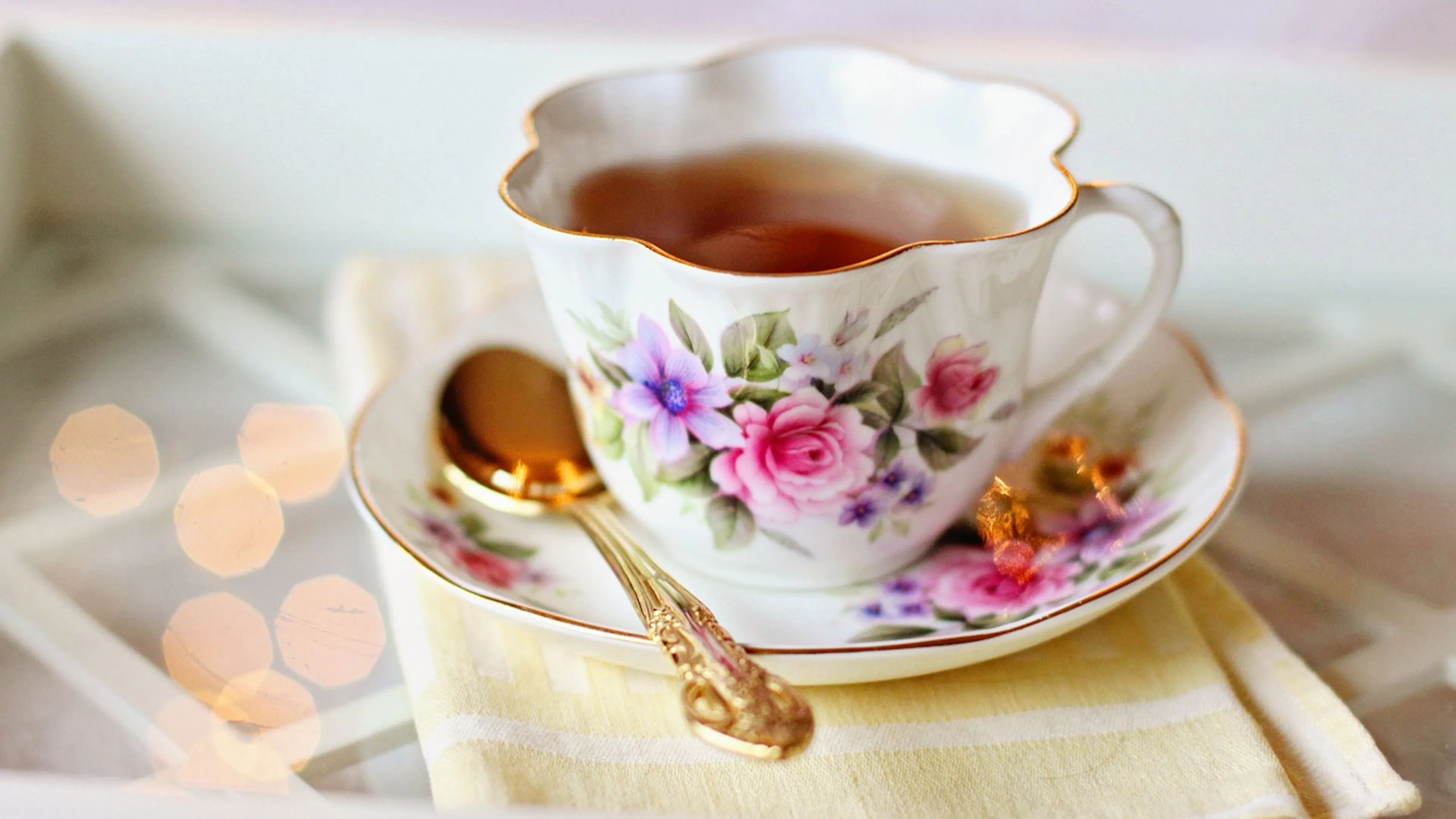A delicate white tea cup painted with small flowers filled with tea on a similar saucer and and next to a gold spoon