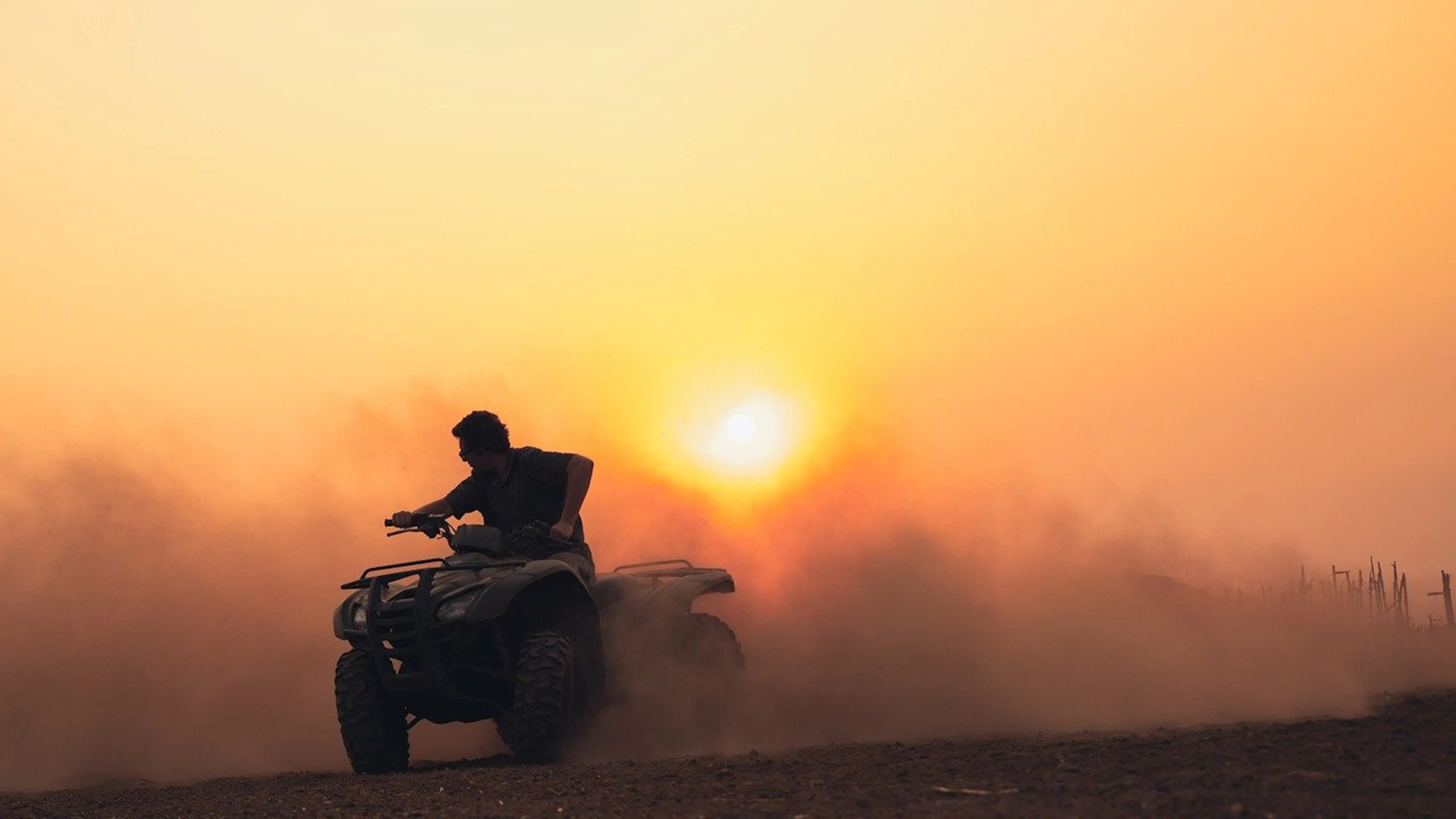 A person driving an ATV through a dusty field kicking up clouds of dark red dust