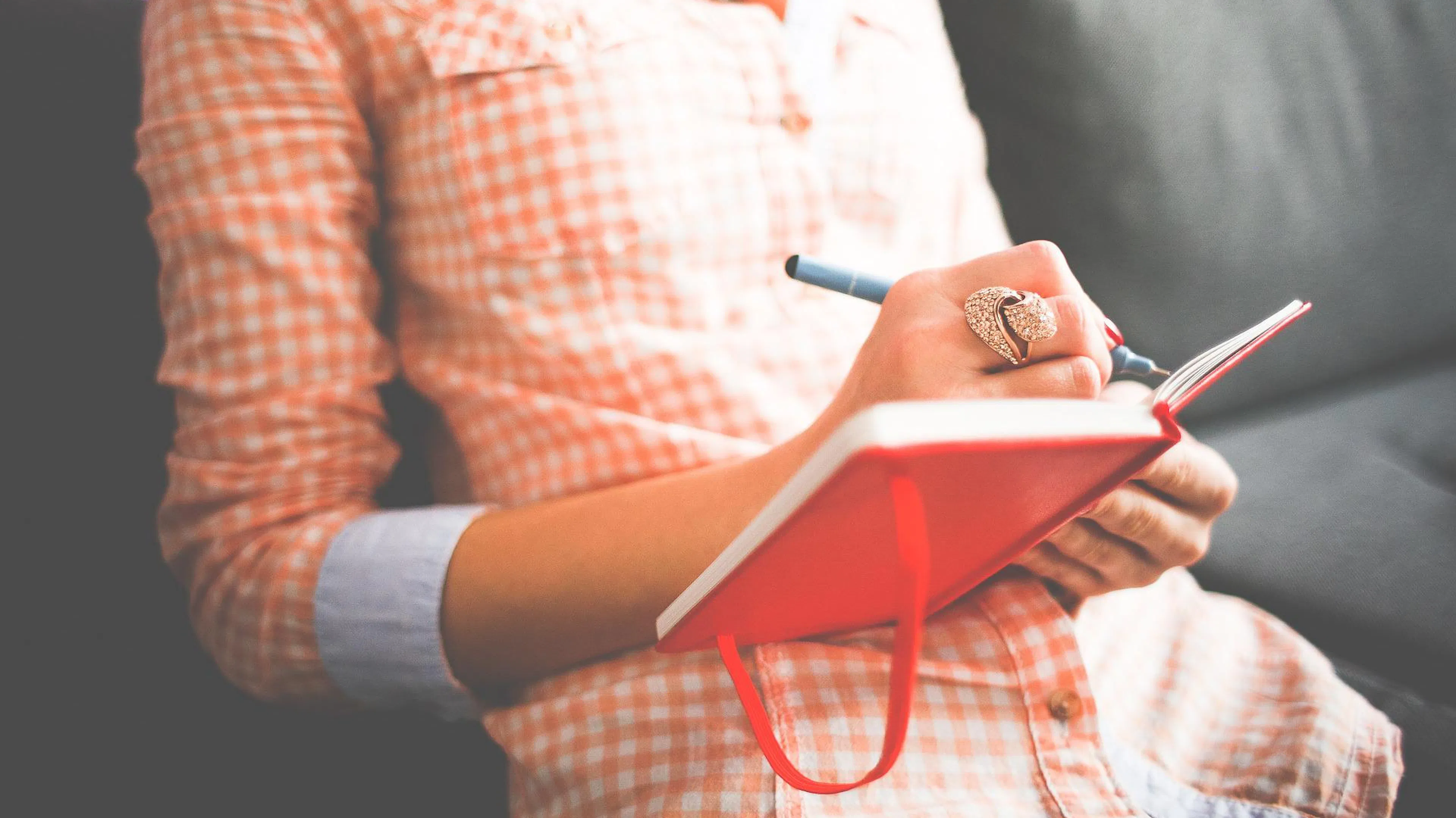 woman in orange checked shirt sitting on couch and writing in red journal