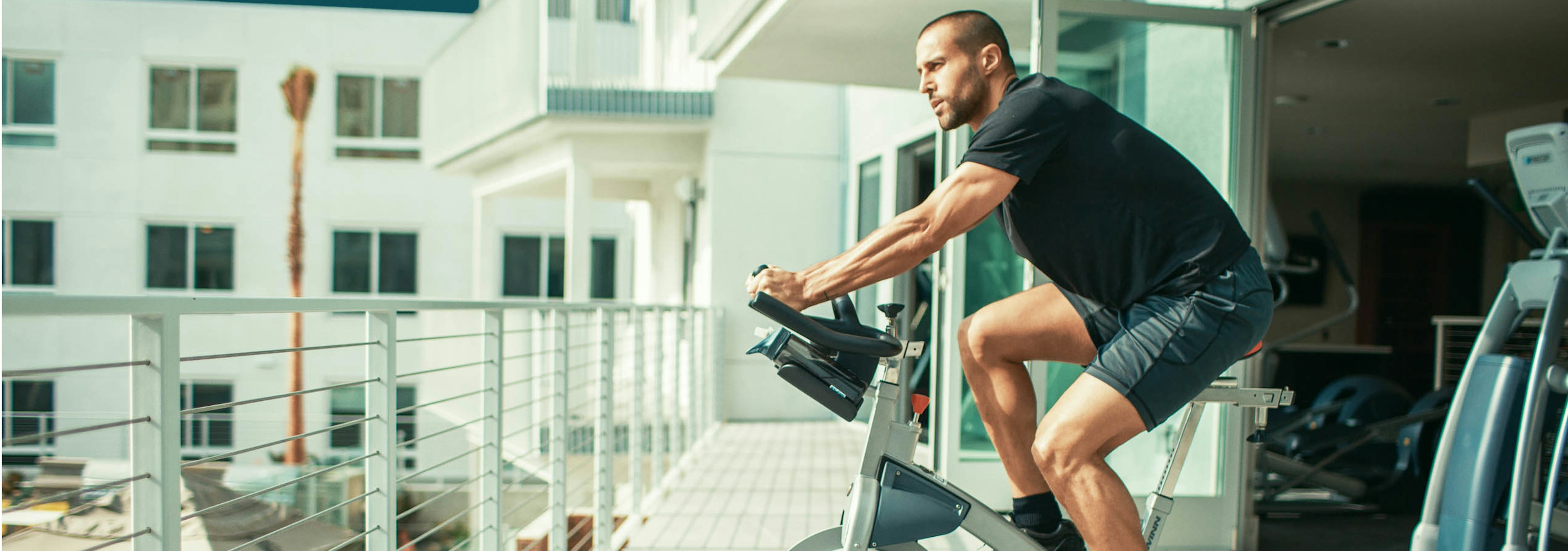 Daytime exterior view of man in shorts on spin bike on yoga deck overlooking the pool area at AMLI Lex on Orange apartments