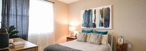 A bedroom at AMLI Dry Creek apartments with gray and blue linens and a dressers with plants and a painting and large window