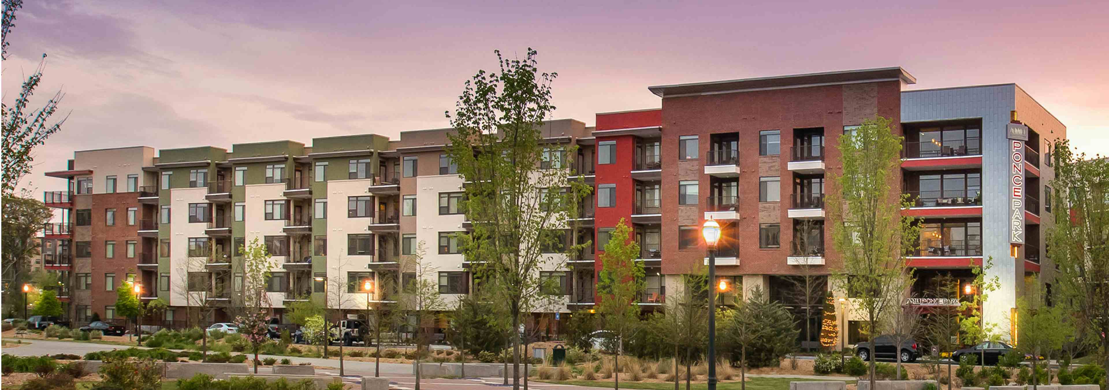 Nighttime exterior view of AMLI Ponce Park brick and stucco apartment building with a tree lined sidewalk and light posts