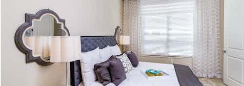 Interior view of AMLI RidgeGate bedroom with king size bed, white duvet and purple pillows facing windows with sheer curtains