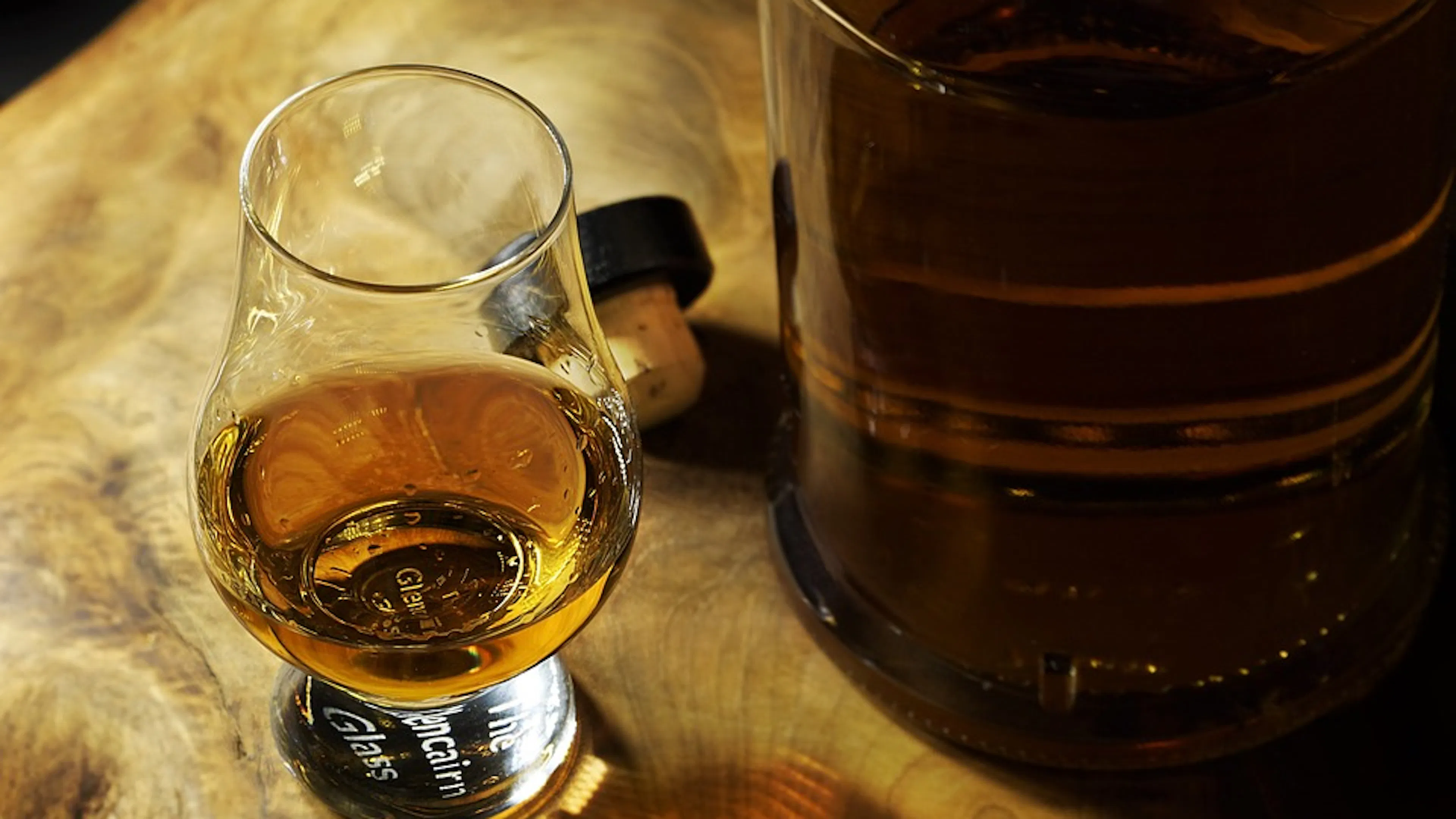 A small glass of bourbon on a wooden table next to a larger glass bottle of the same dark bourbon