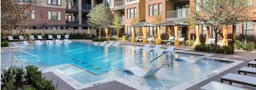 View of pool at AMLI West Plano facing the building facade with in water lounge chairs and surrounding vibrant trees
