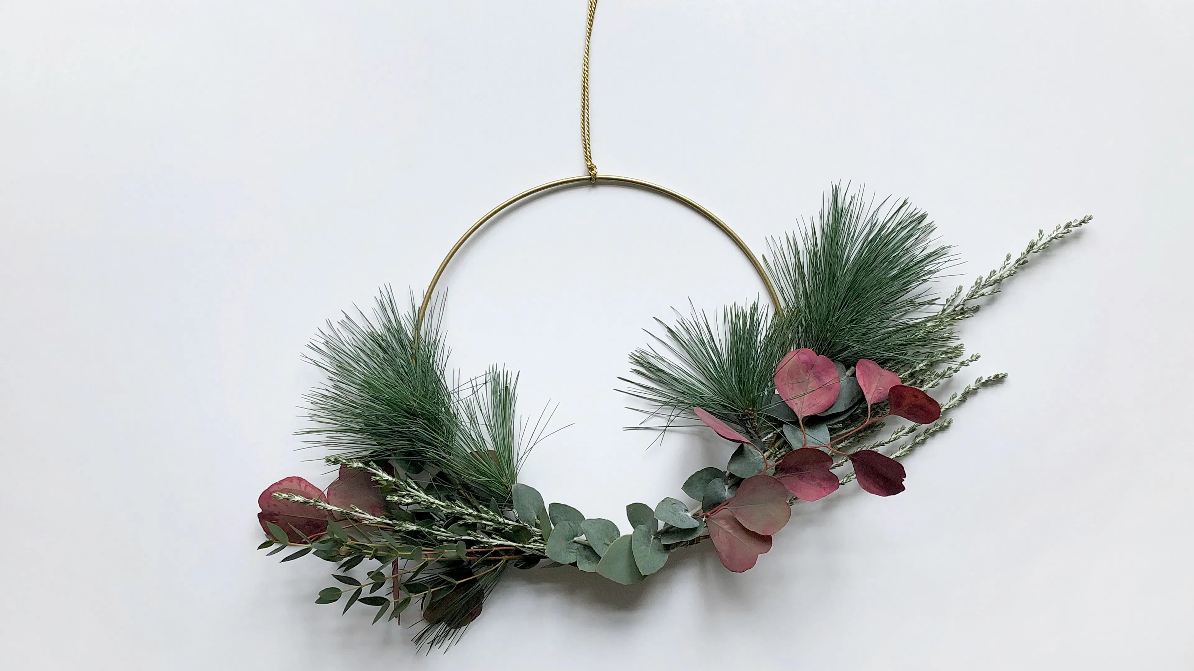 A thin gold wire circle with pine boughs and eucalyptus leaves tied onto it with twine and hung by a thin piece of ribbon