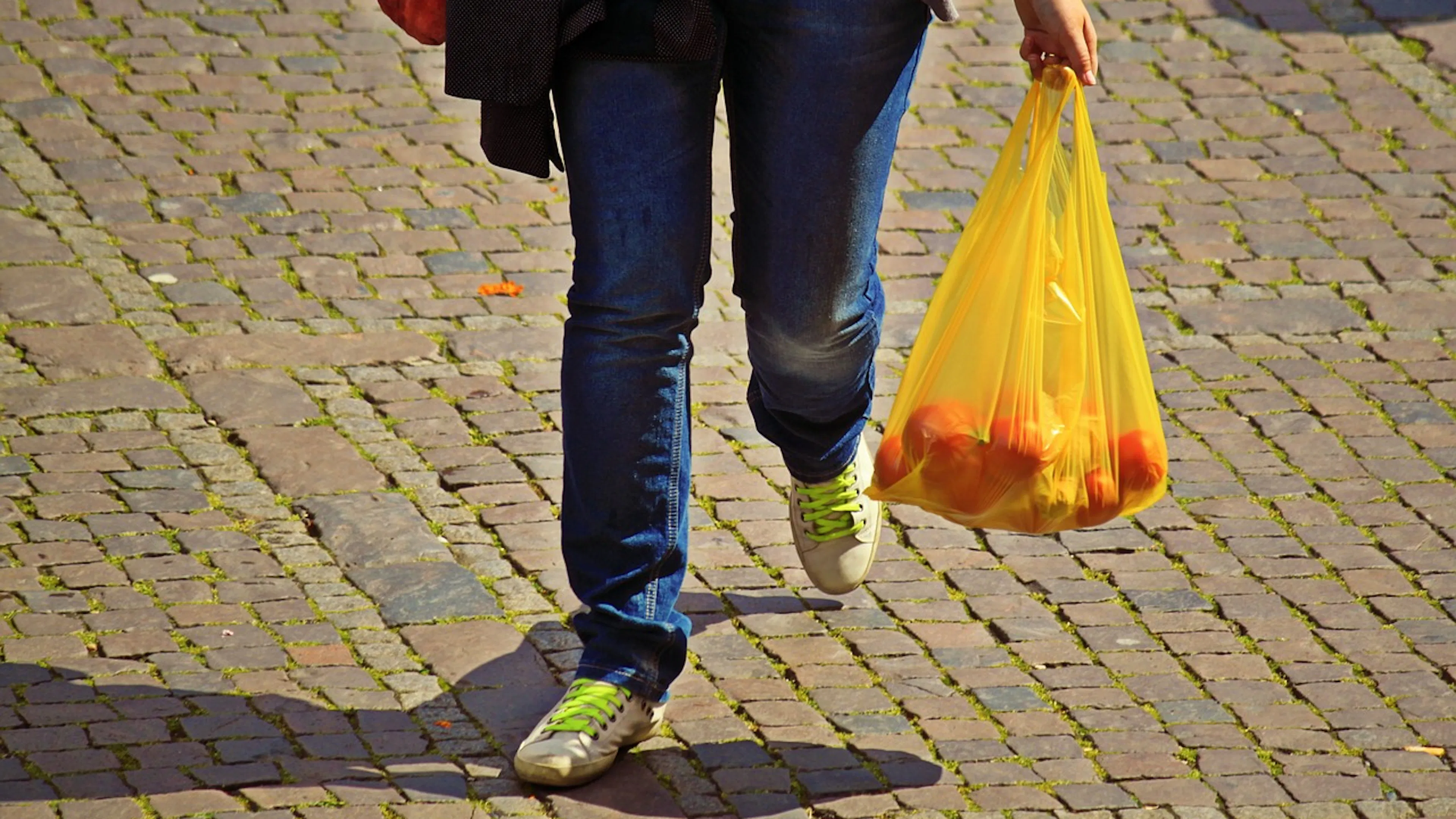 Picture of a walking person from the waist down holding a yellow plastic bag of oranges 