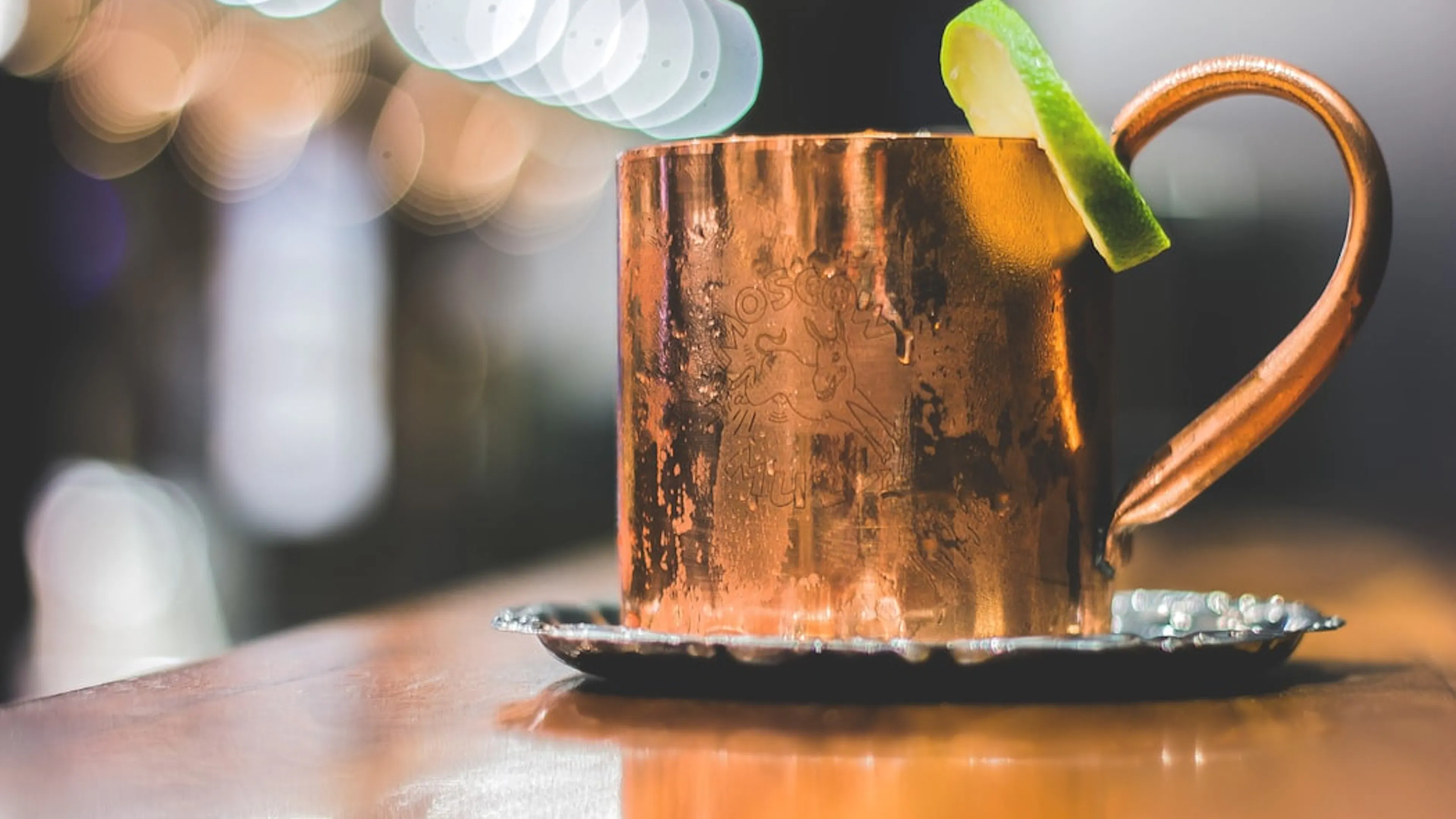 A copper Moscow Mule cup with a lime wedge garnishing the rim on a wooden bar counter 
