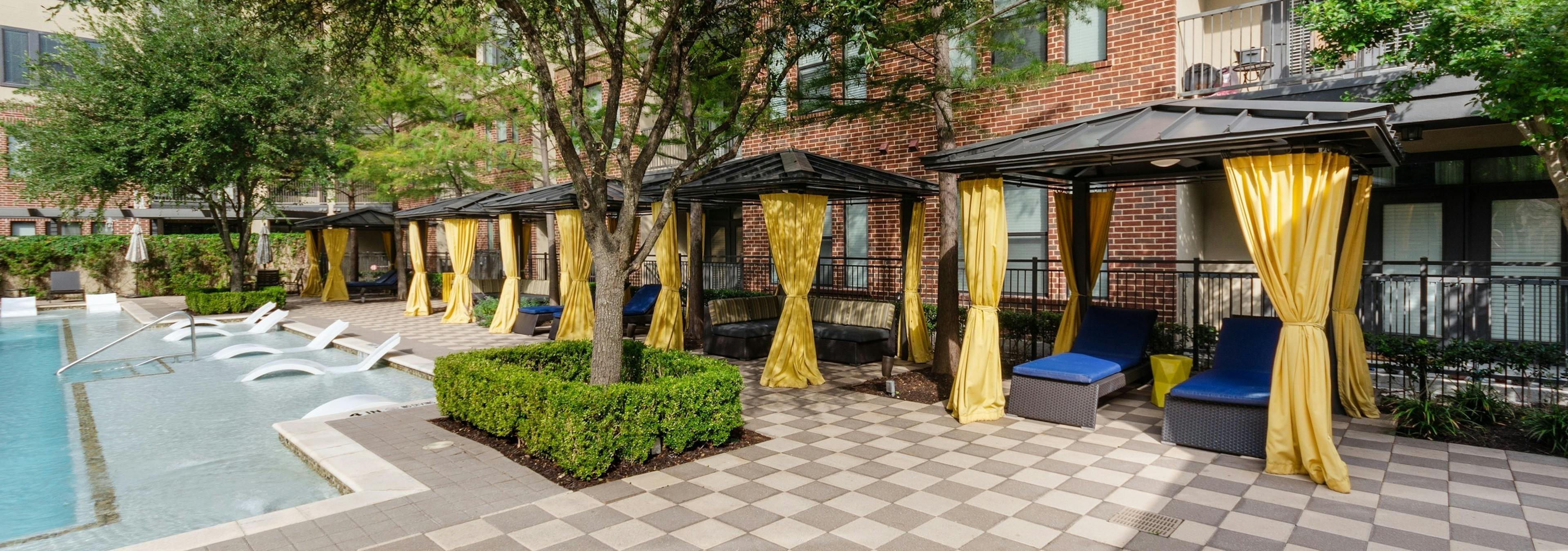 Daytime view of AMLI West Plano resident cabanas with yellow privacy curtains and blue lounge seating facing the pool