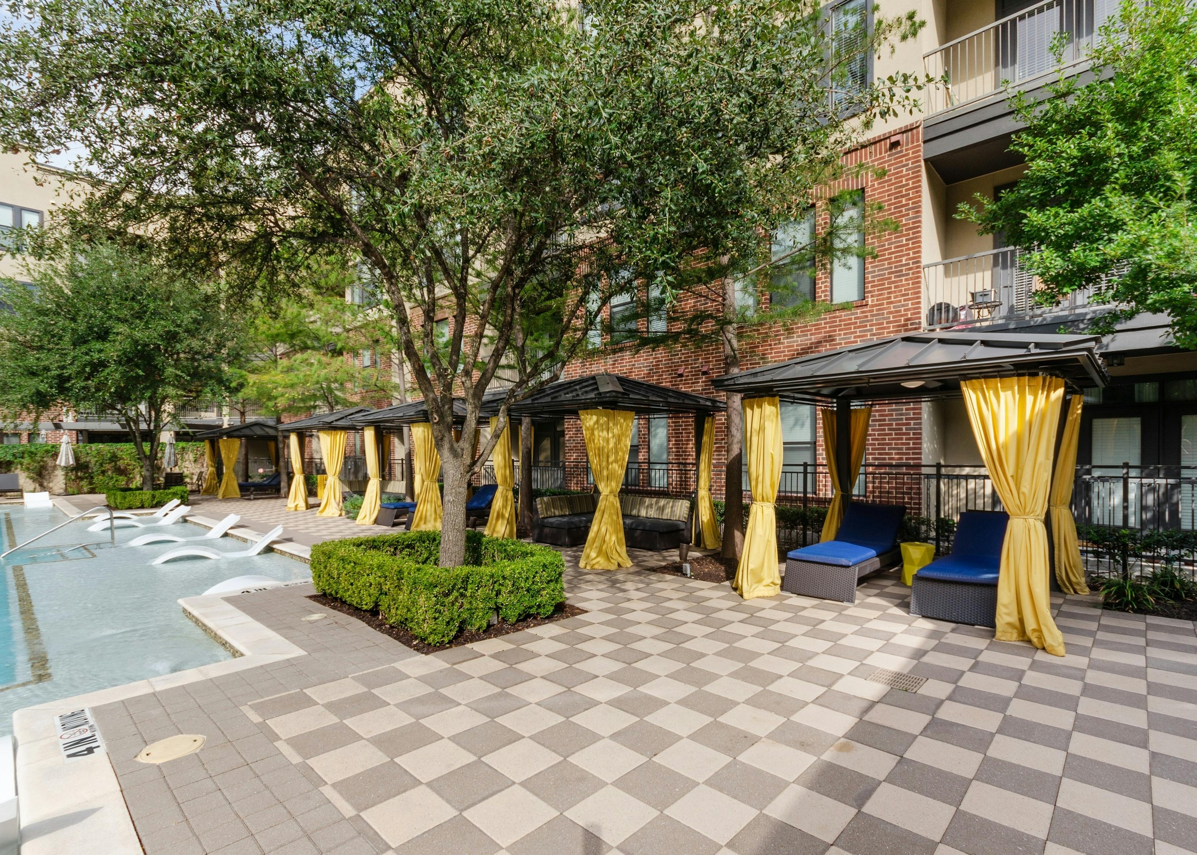 Daytime view of AMLI West Plano resident cabanas with yellow privacy curtains and blue lounge seating facing the pool