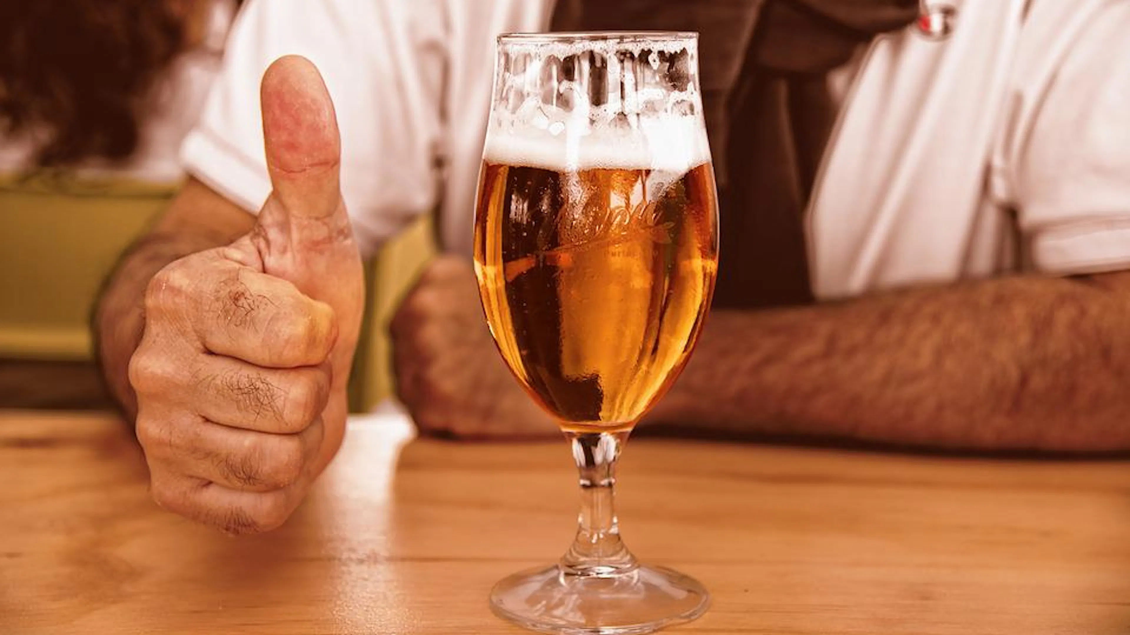 A man sitting at a bar giving a thumbs up beside a glass full of beer