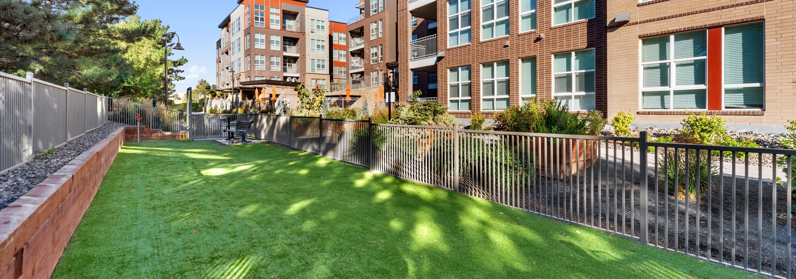 A view at the paw park at AMLI Dry Creek apartments with green grass and bench at far with  surrounding fence and brick building in rear